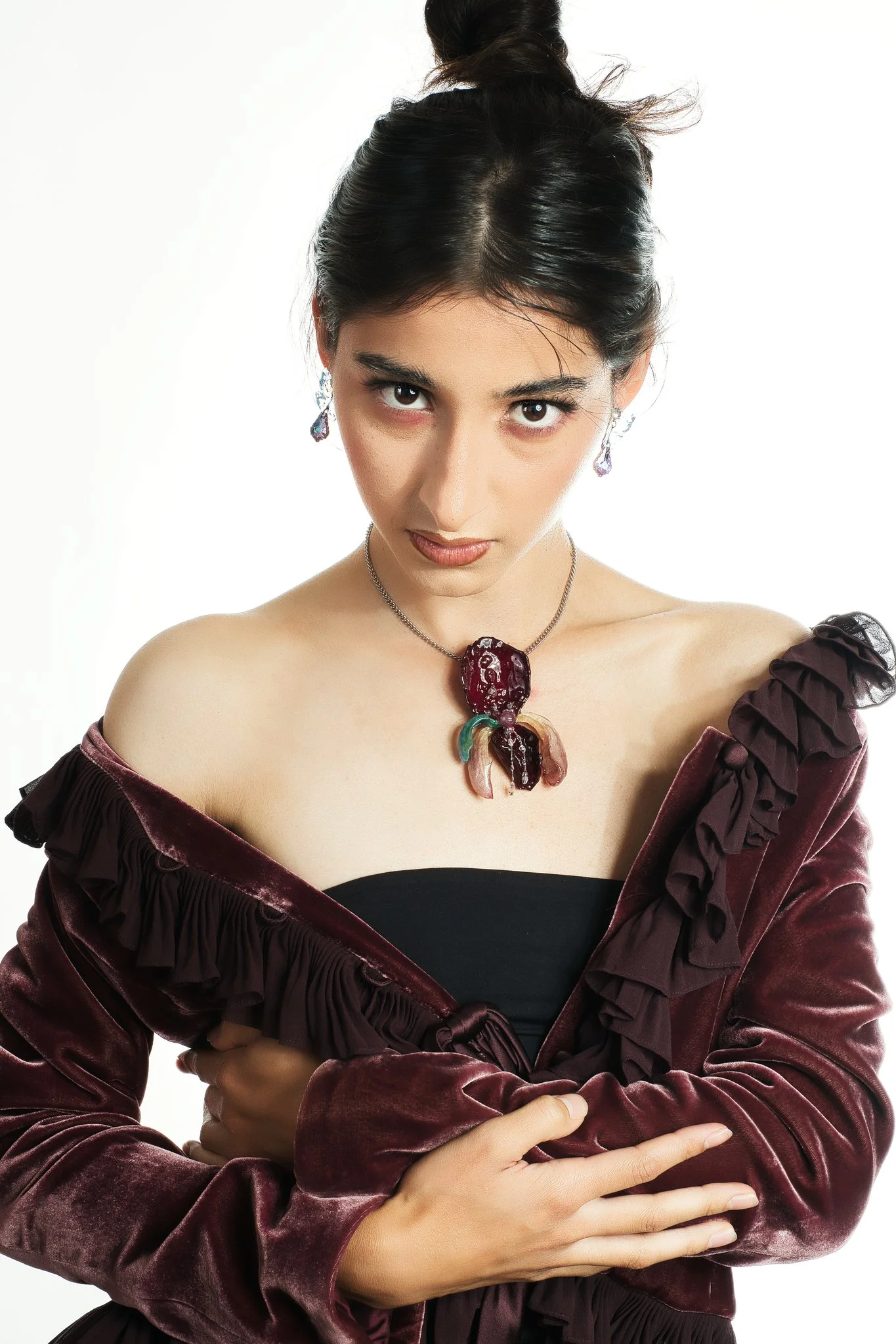 A woman with dark hair styled in an updo, wearing earrings, a necklace with a bold, dark red pendant, and a velvet burgundy off-shoulder top, posing against a white background.
