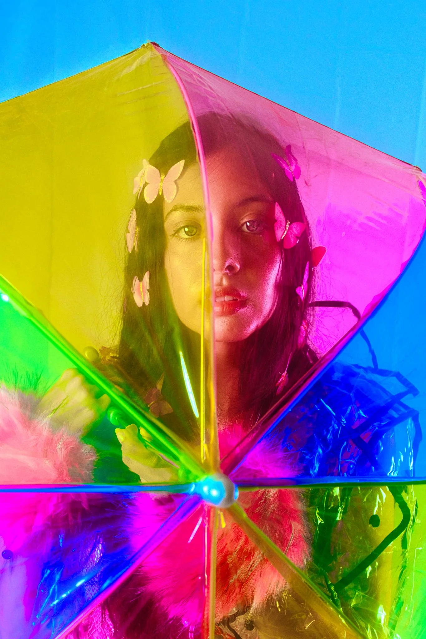 A woman holding a multicolored, translucent umbrella with butterfly patterns, with feathers and bright neon lighting.