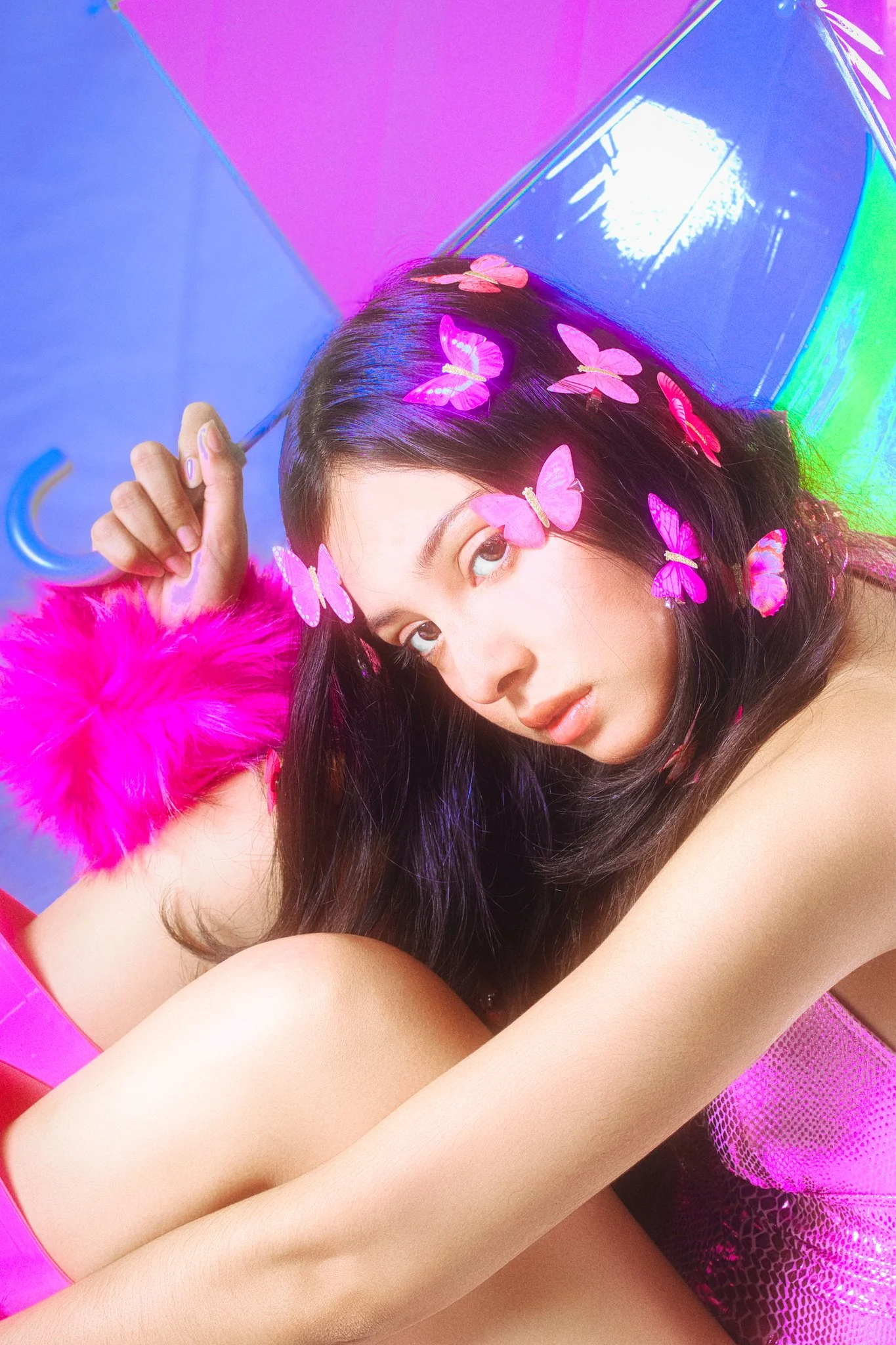 A young woman with dark flowing hair decorated with pink butterflies, lying on a colorful pink and purple surface with an iridescent box nearby.