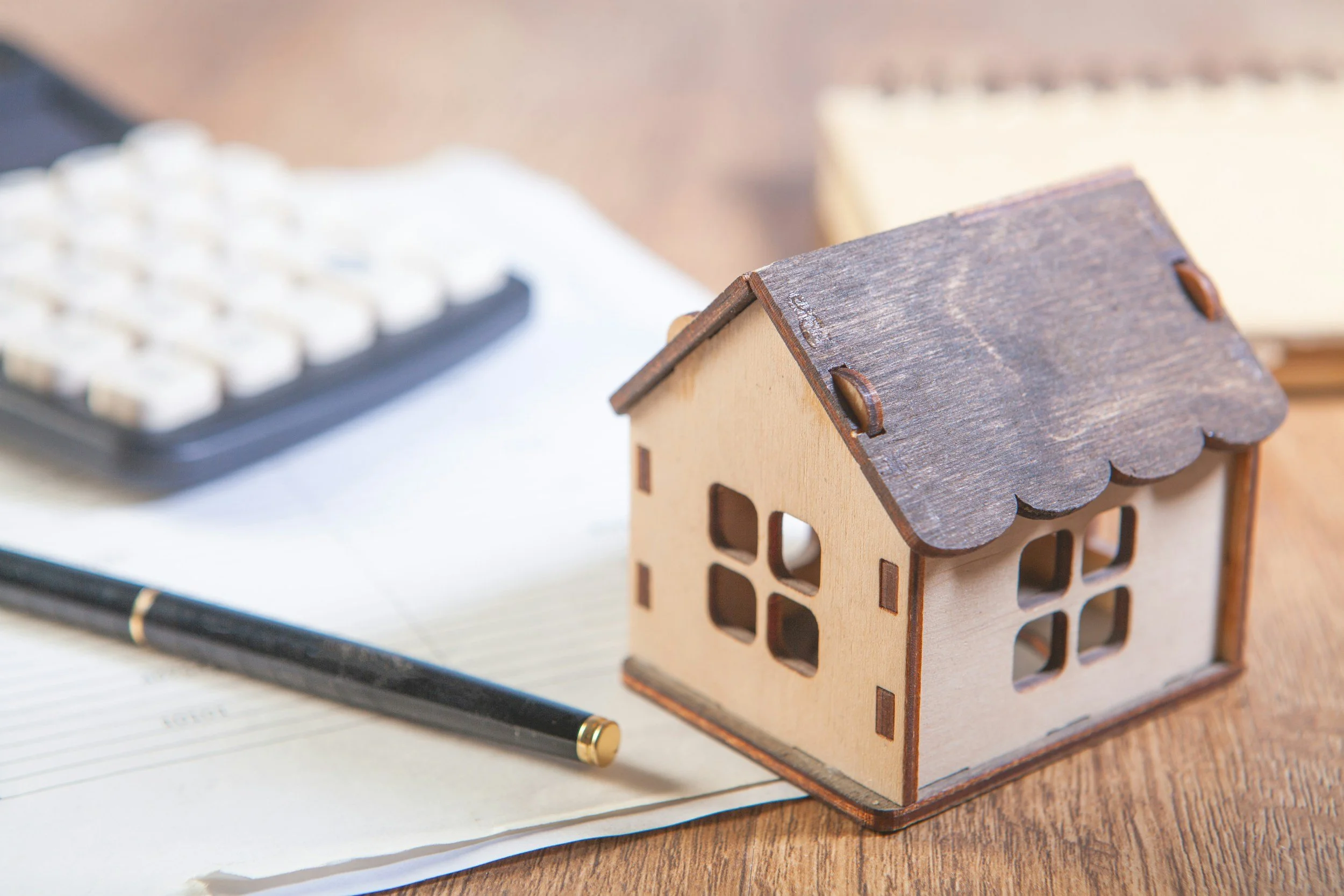 A small wooden house model placed on a table, next to a pen and some papers with a calculator in the background.