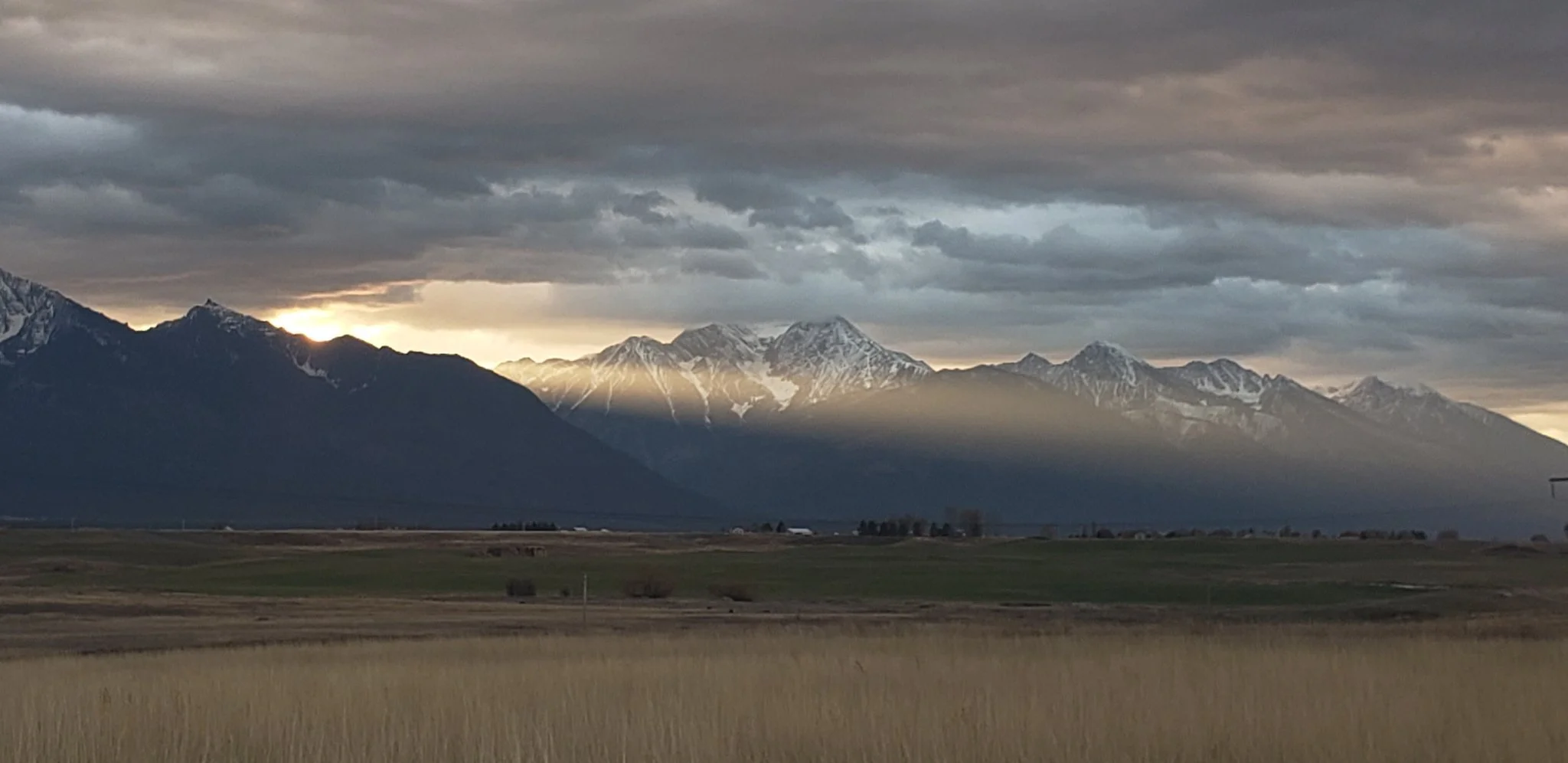 A scenic view of snow-capped mountains under a cloudy sky with sunlight breaking through, viewed from a grassy field.