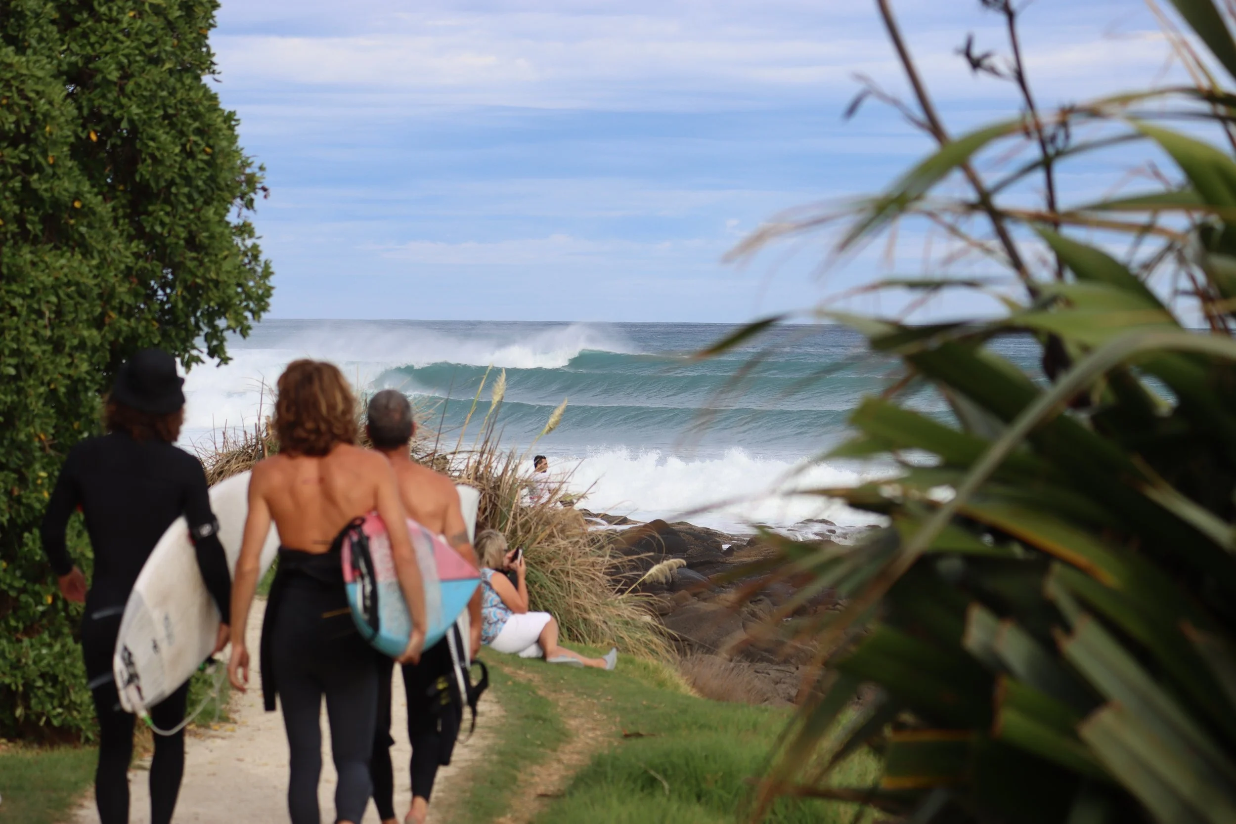 People sitting and walking on a beachside path, at Whale Bay, Raglan. Large waves crashing on the shore, green bushes, and an overcast sky in the background.