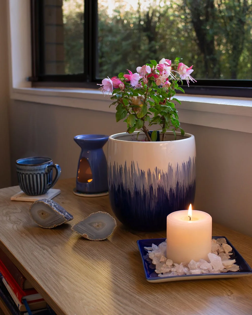 A decorative arrangement on a wooden table near a window, featuring a large potted pink flowering plant, a blue and white candle, two geode slices, a blue ceramic cup, a blue ceramic oil warmer, and a lit candle on a square blue dish surrounded by white stones.