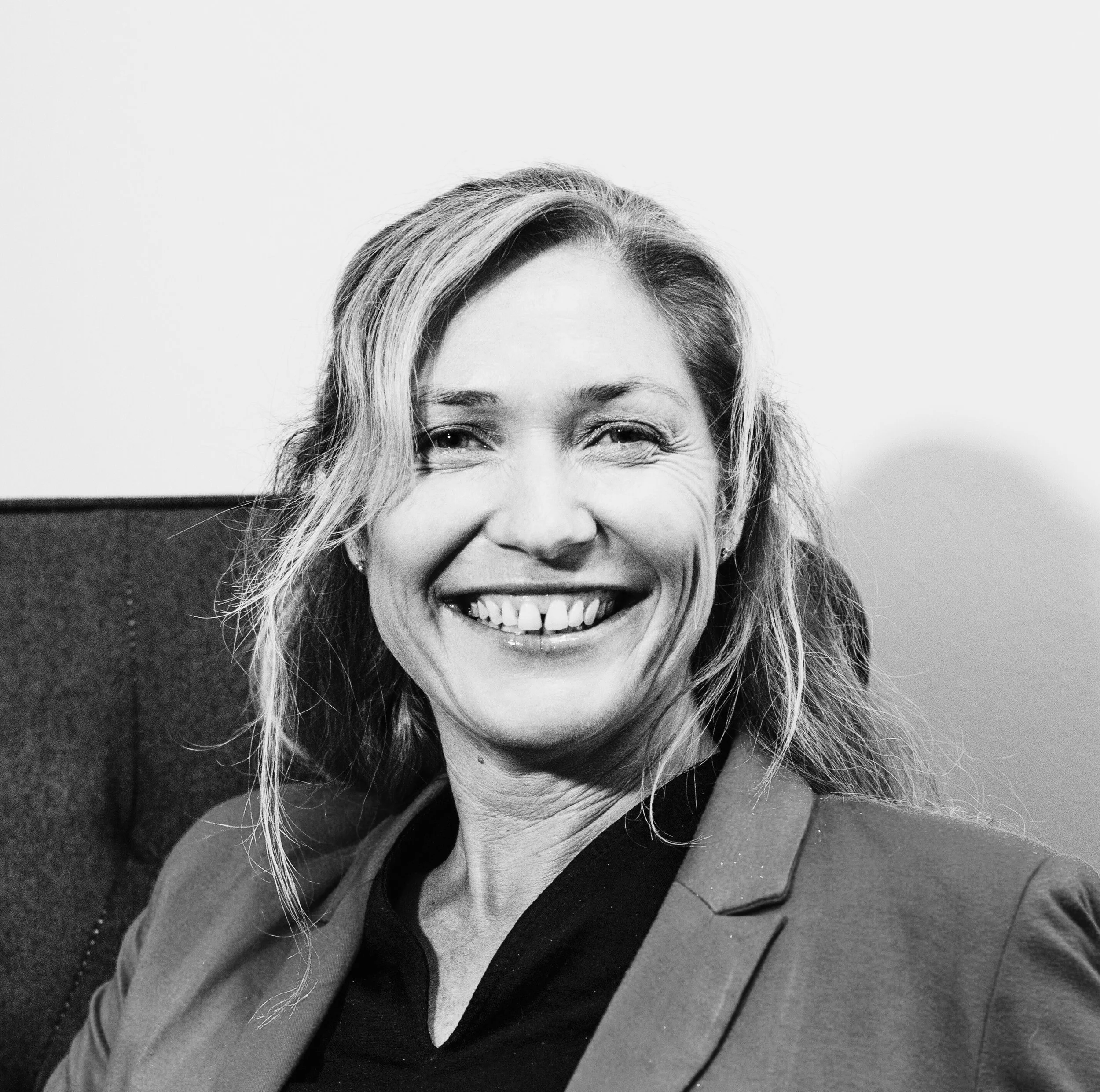 A smiling woman with wavy hair wearing a blazer and sitting on a chair.