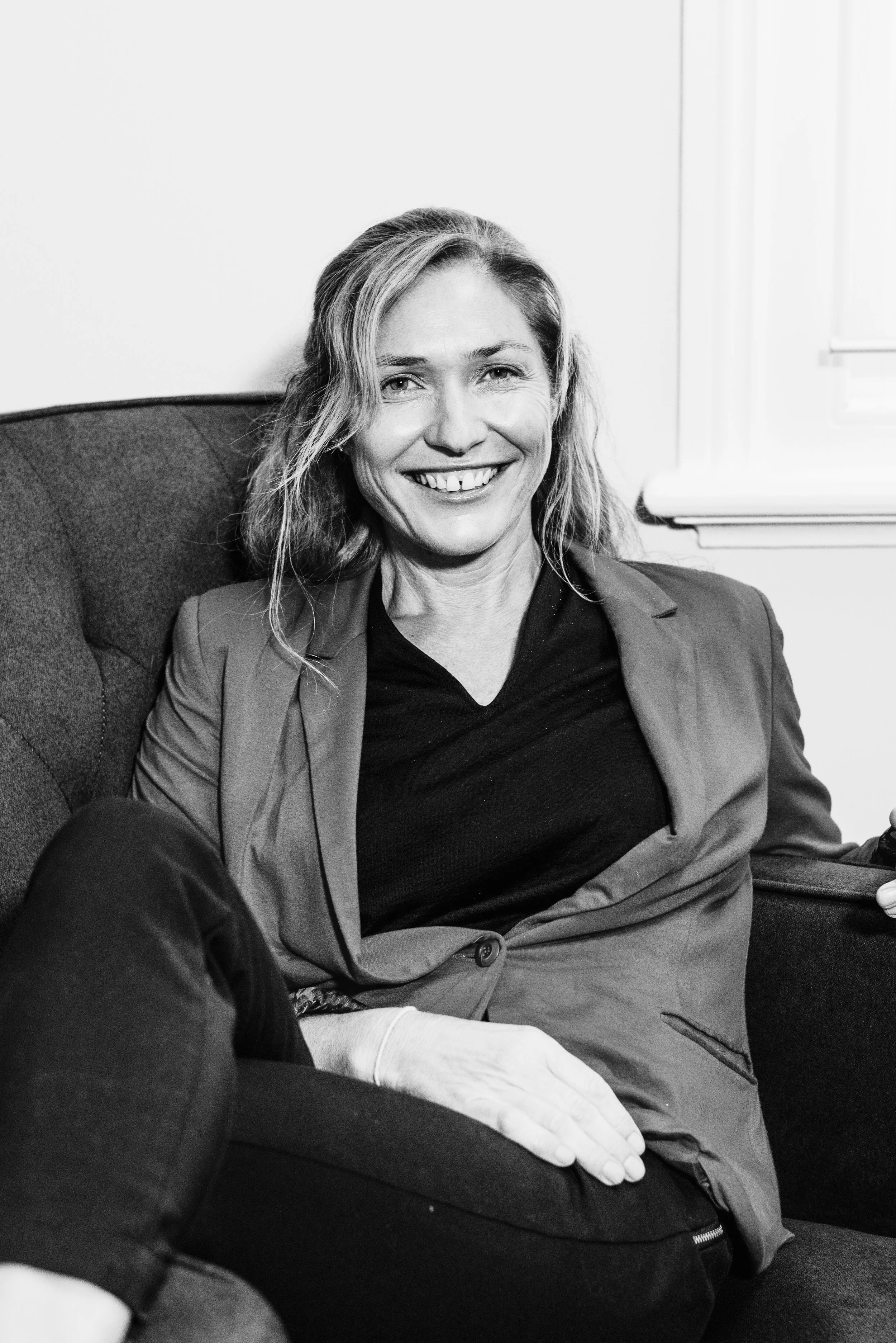 A woman with wavy hair, wearing a blazer and a black top, sitting on a sofa, smiling at the camera, in a black-and-white photo.
