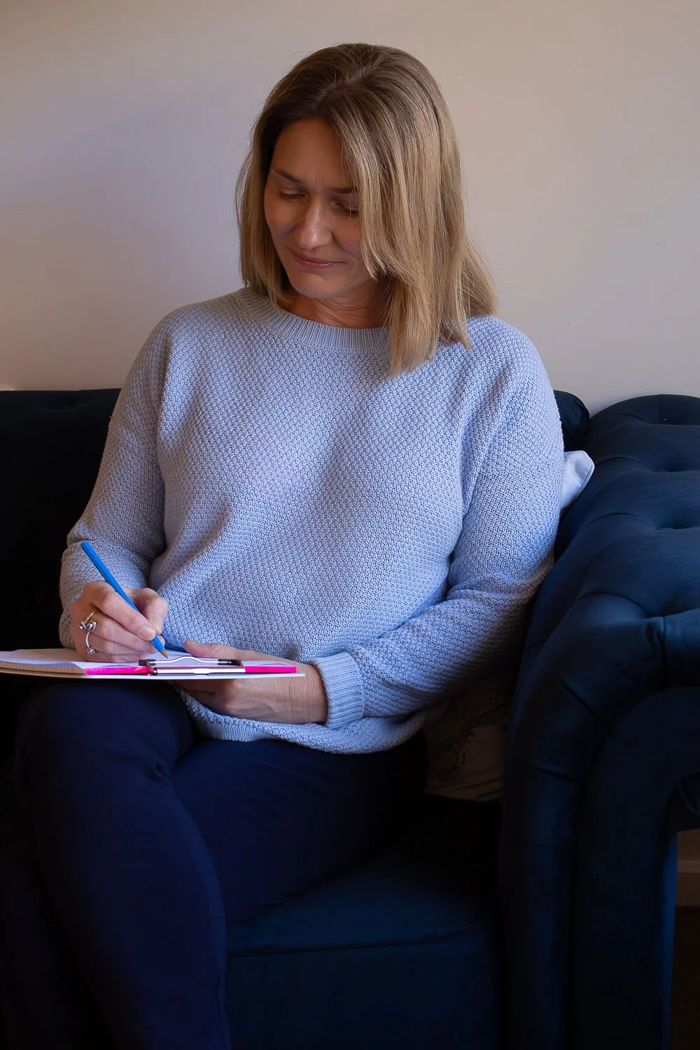 A woman with shoulder-length blonde hair wearing a light blue jumper sitting on a black couch, writing in a notebook with a blue pen.