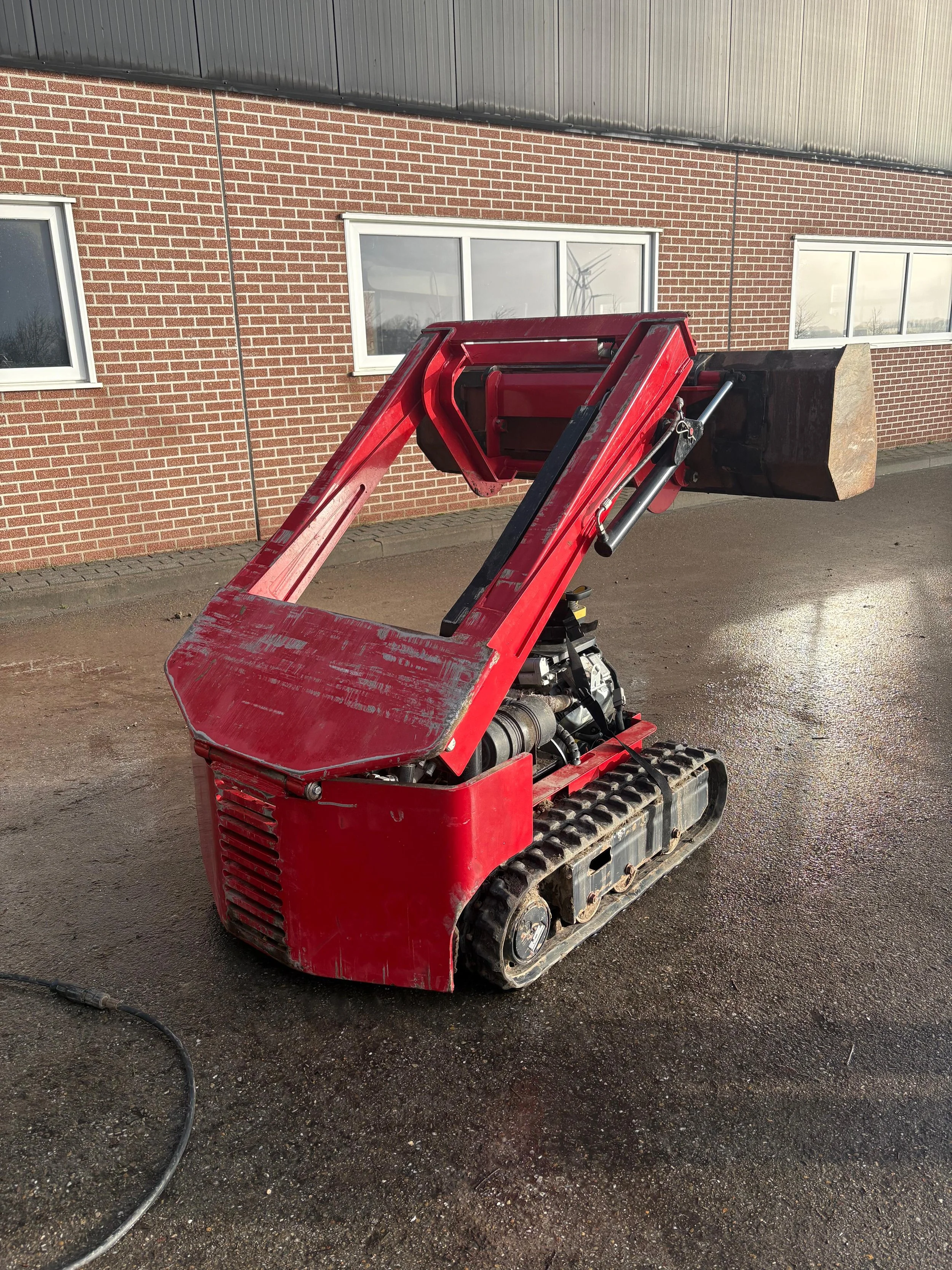 Een kleine rode graafmachine met tracks, geparkeerd op een zandige ondergrond voor een gebouw met bakstenen en metalen gevelpanelen.