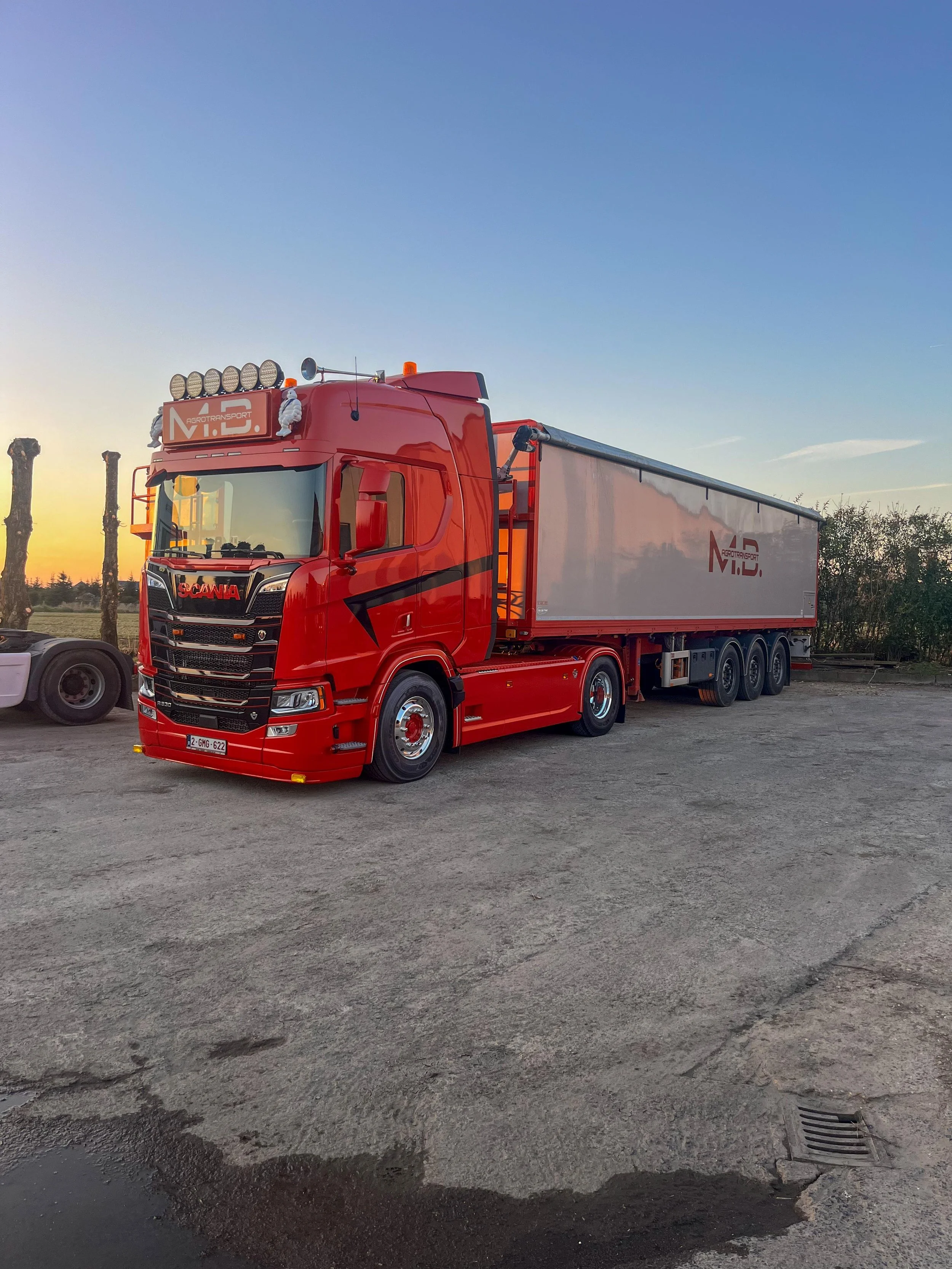 Een grote rode vrachtwagen met een witte trailer, geparkeerd op een onverharde parkeerplaats bij zonsondergang.