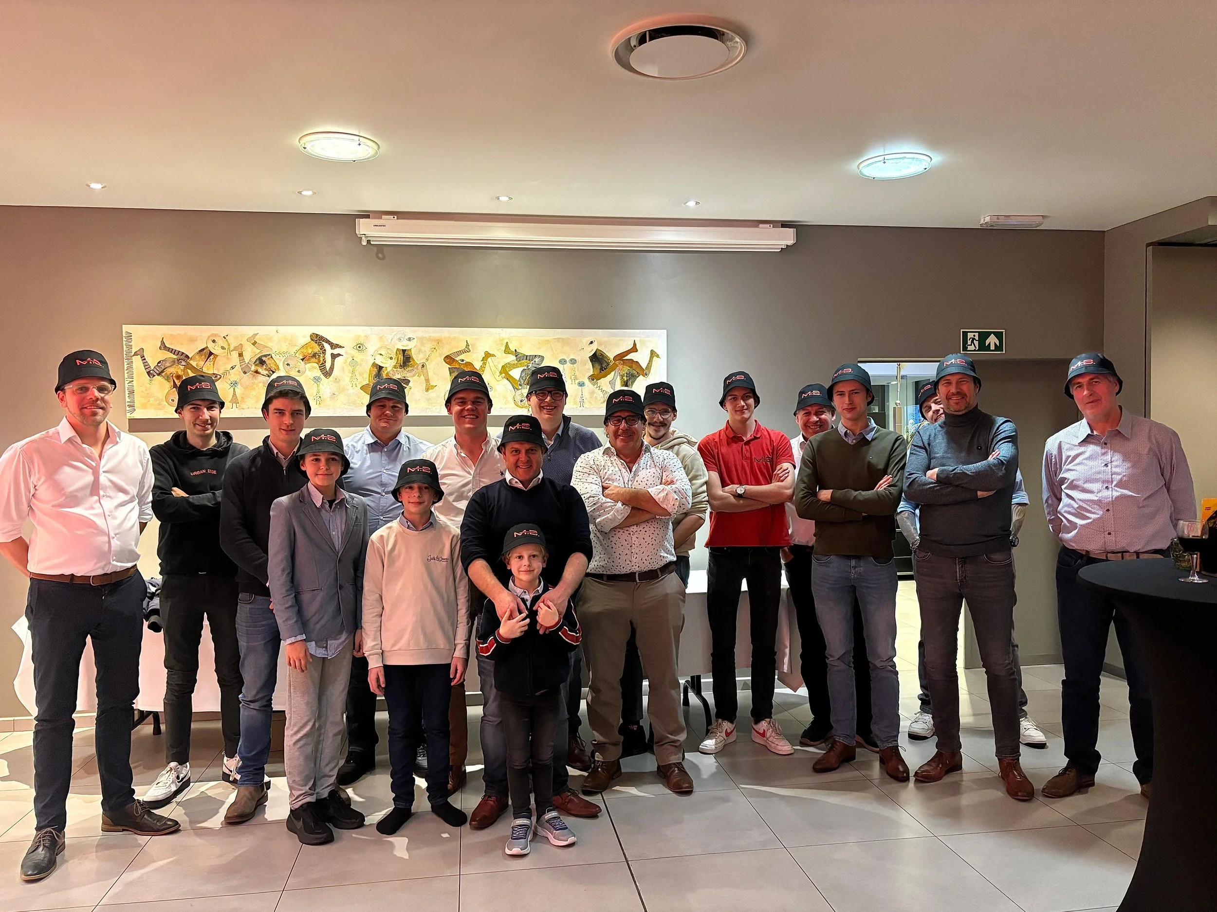 Groep mensen poseert voor een foto in een binnenruimte, velen dragen zwarte petten met een rood logo.