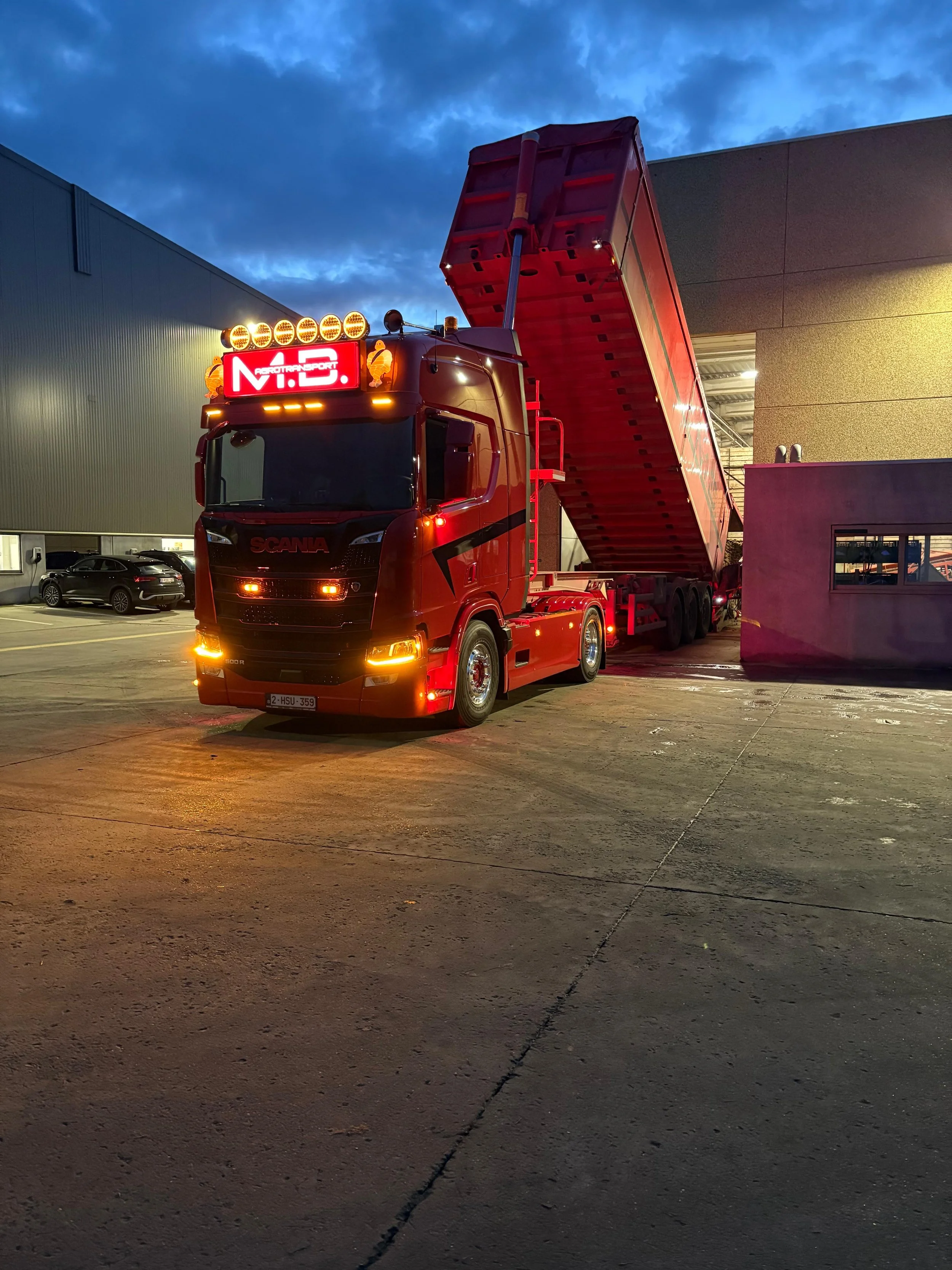 Een rode vrachtwagen met een open laadbak, geparkeerd op een parkeerplaats bij schemerdonker, naast een industrieel gebouw met een kleine ramen en een een zwarte auto in de achtergrond.