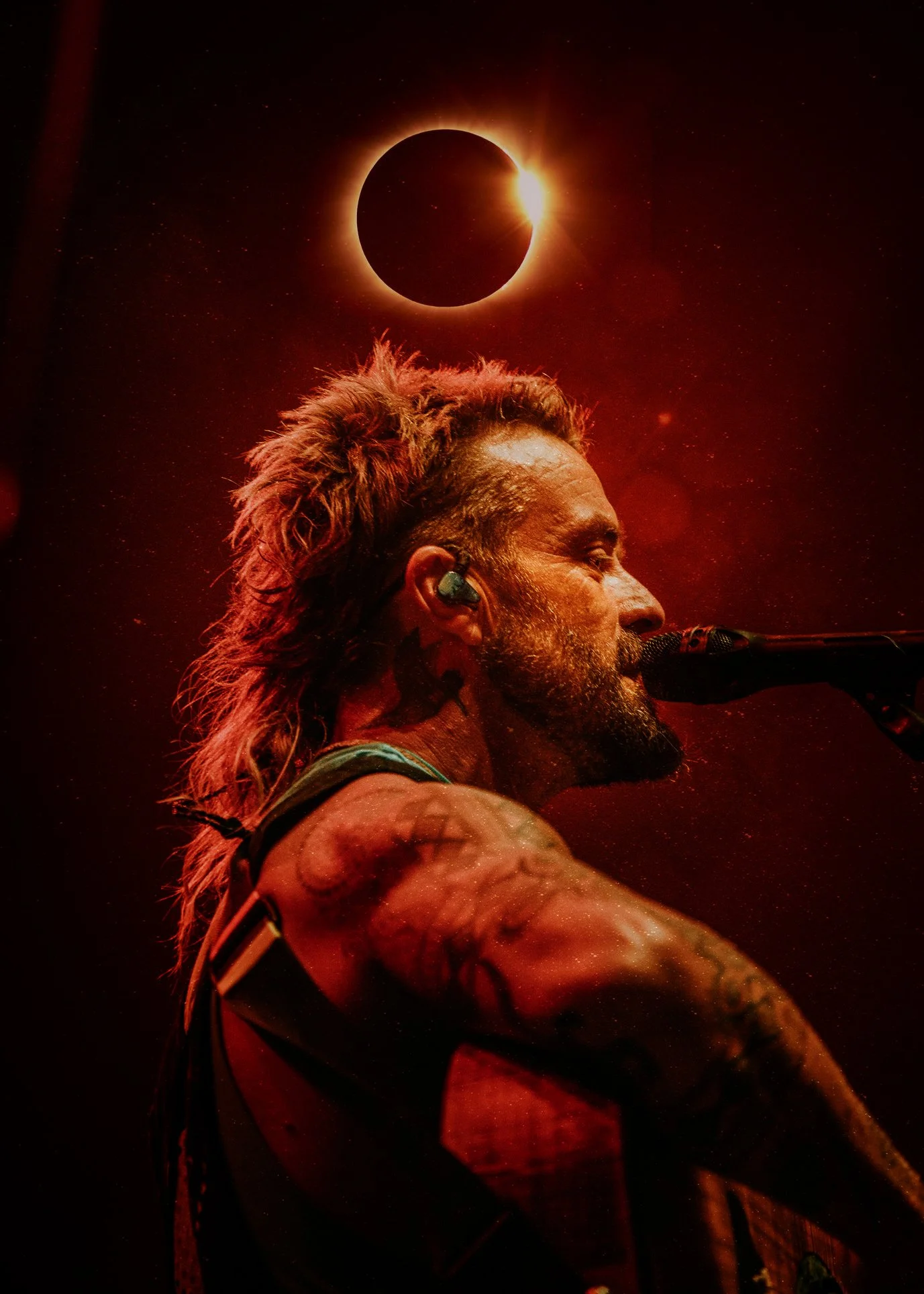 A male musician with tattoos and long hair singing into a microphone during a performance, with a solar eclipse and a bright star in the background.