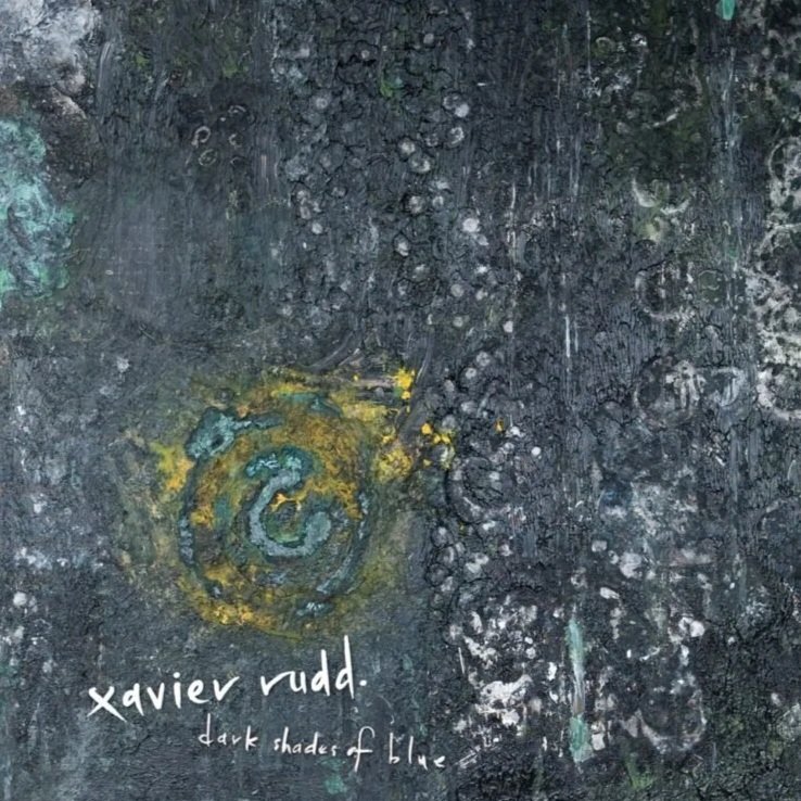 Close-up of a textured, dark abstract artwork with yellow and blue painted swirls, and handwritten white text that reads 'Xavier Rudd' and 'dark shades of blue'.