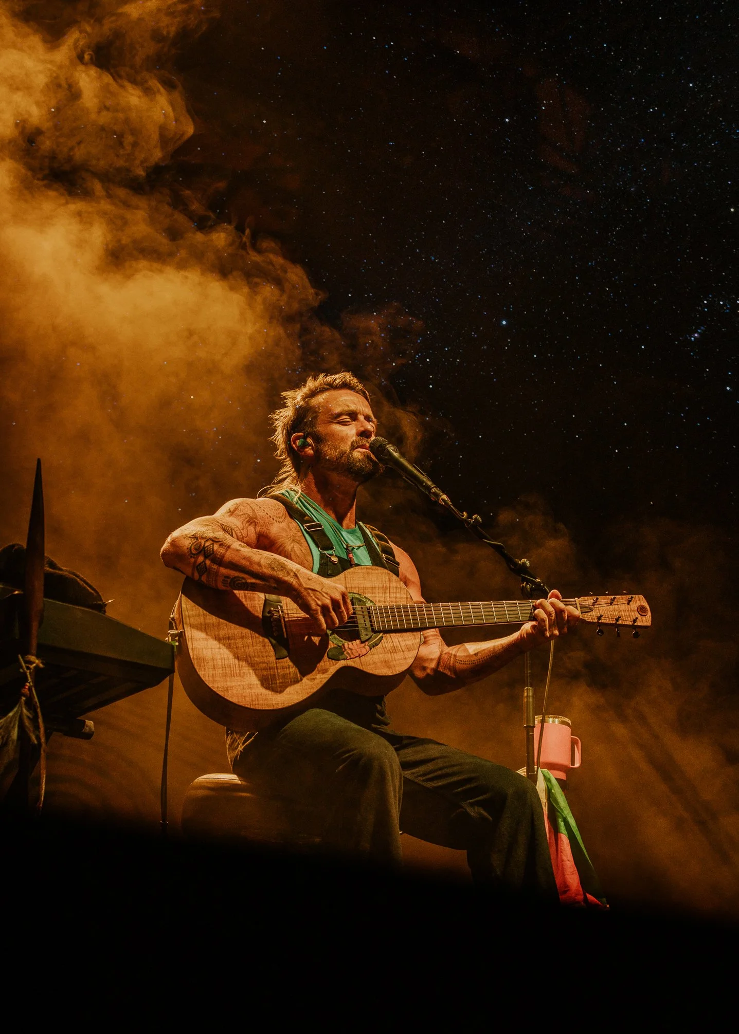 A male musician with tattoos playing an acoustic guitar and singing into a microphone during a performance, with orange stage lighting, smoke, and a dark night sky filled with stars in the background.