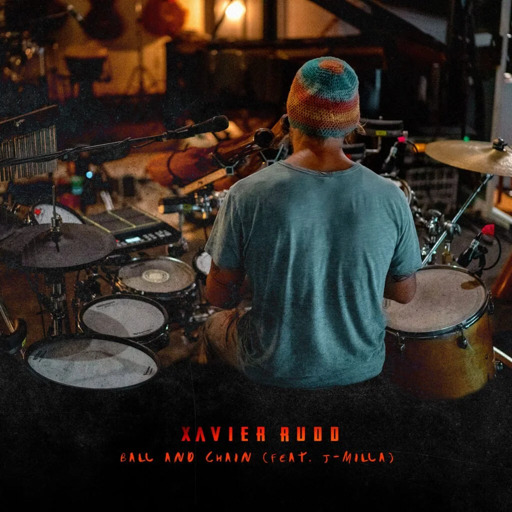 A person wearing a colorful knit hat and a gray t-shirt is playing drums in a dimly lit music studio or venue, with musical equipment and instruments visible in the background.
