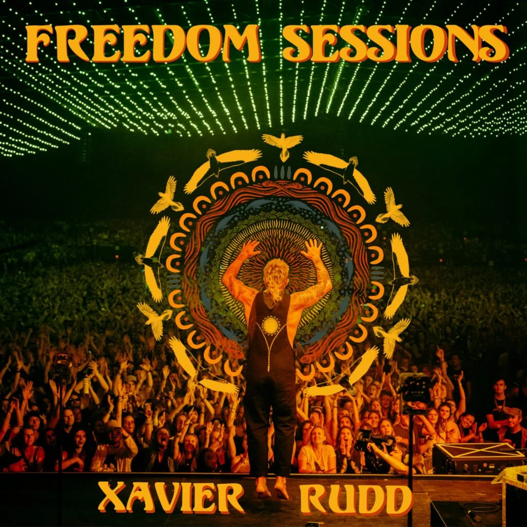A large crowd attending a concert or event, with a person on stage raising their hands. The stage backdrop features an artistic design with concentric circles, birds, and dynamic lines. The words "FREEDOM SESSIONS" are displayed at the top, and "XAVI