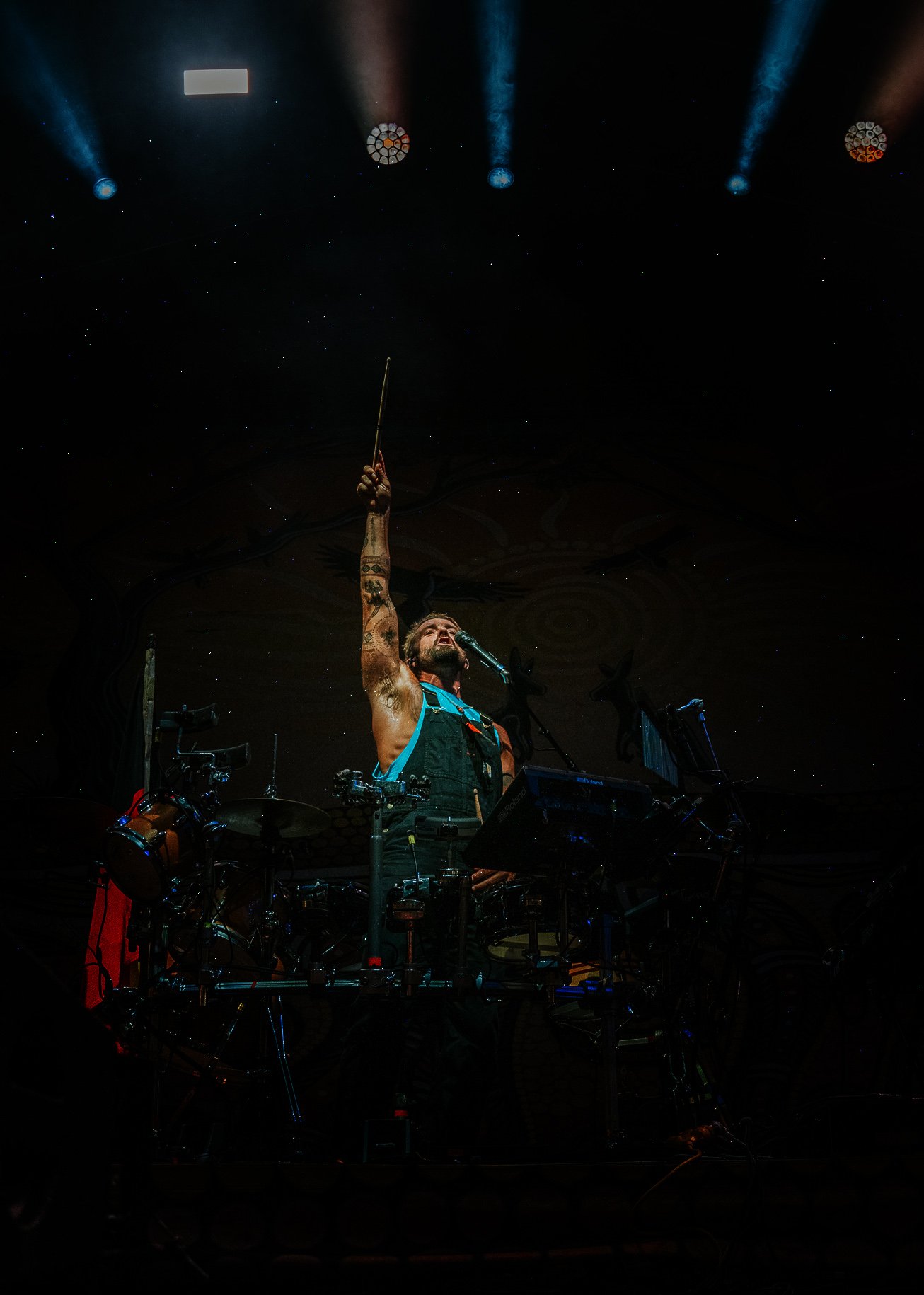 A performer on stage raising a baton, surrounded by a drum set and electronic music equipment, with stage lights overhead.
