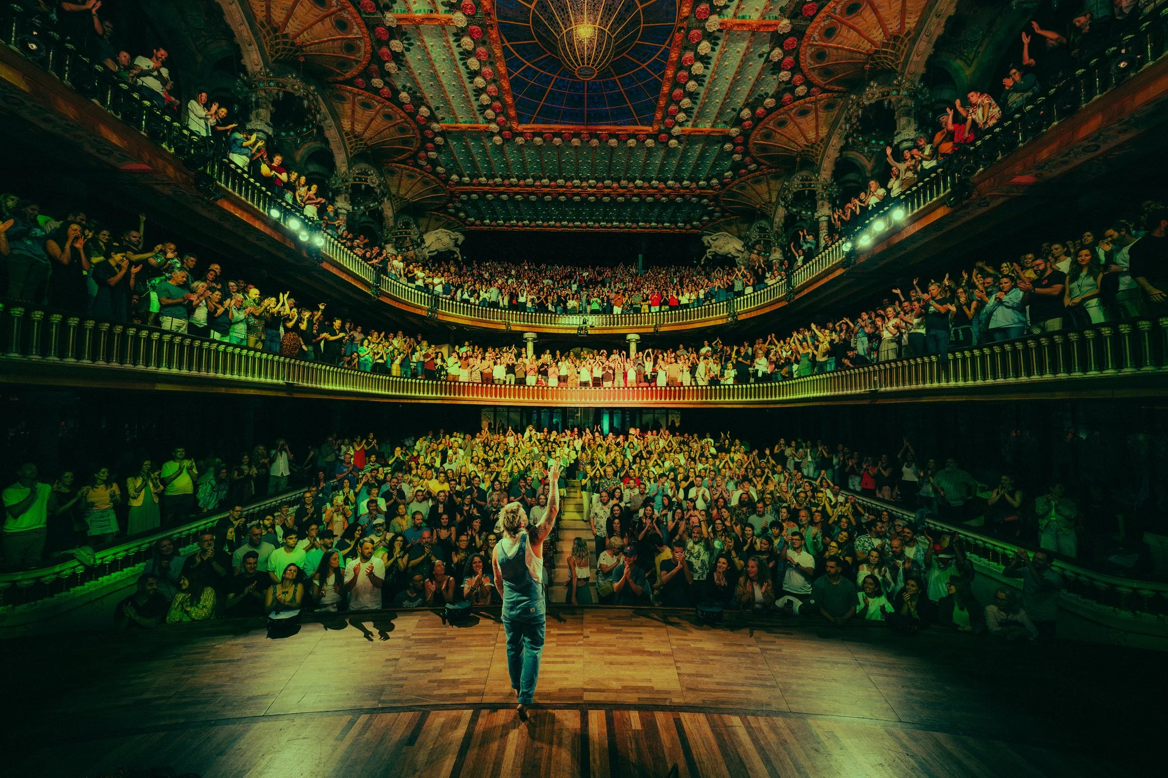 A performer stands on stage in front of a large, packed theater with multiple balconies filled with audience members clapping and cheering.