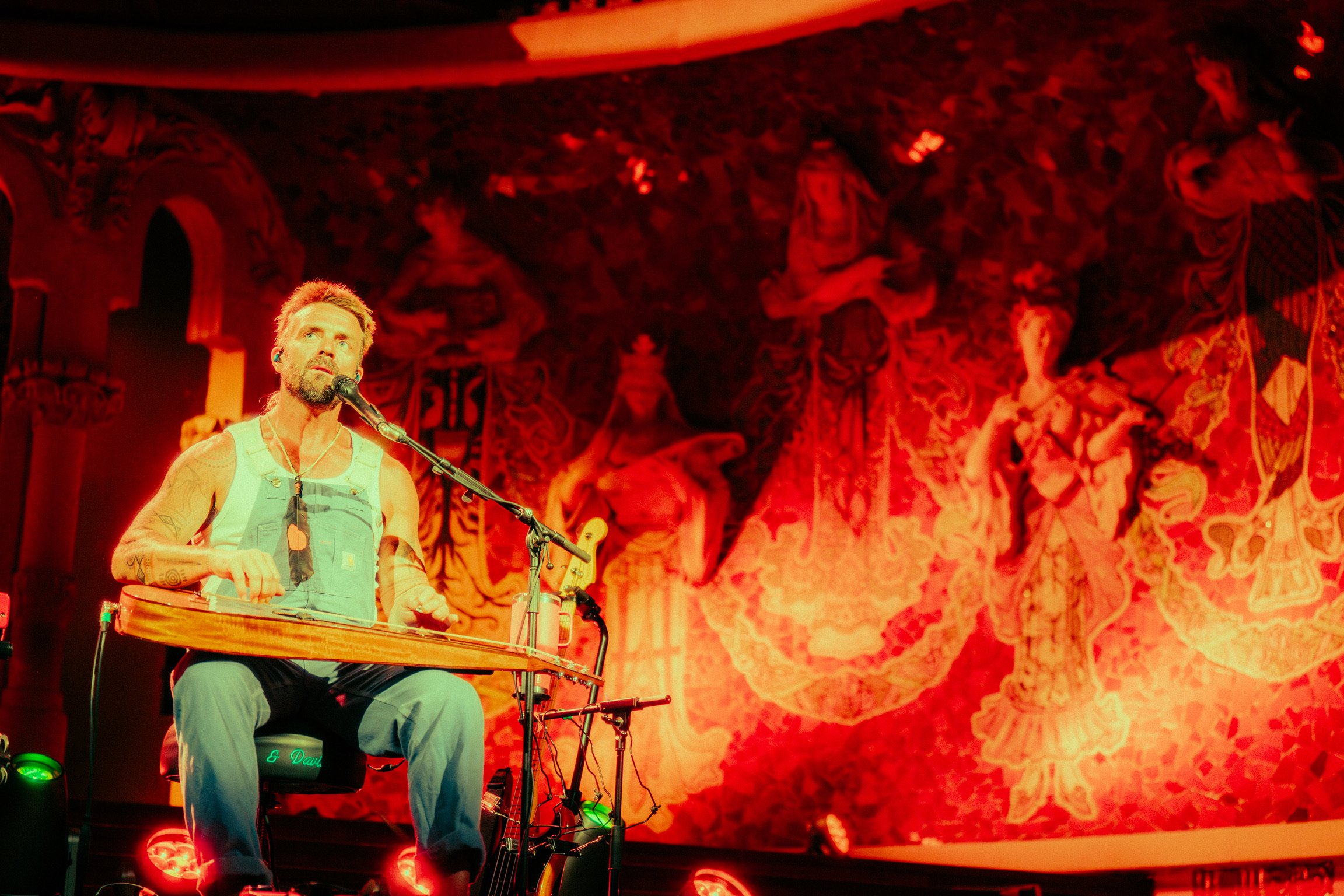 A man with a beard and tattooed arms, wearing overalls and a white tank top, is sitting and playing a lap steel guitar on stage. He is singing into a microphone in front of a red ornate mural backdrop.