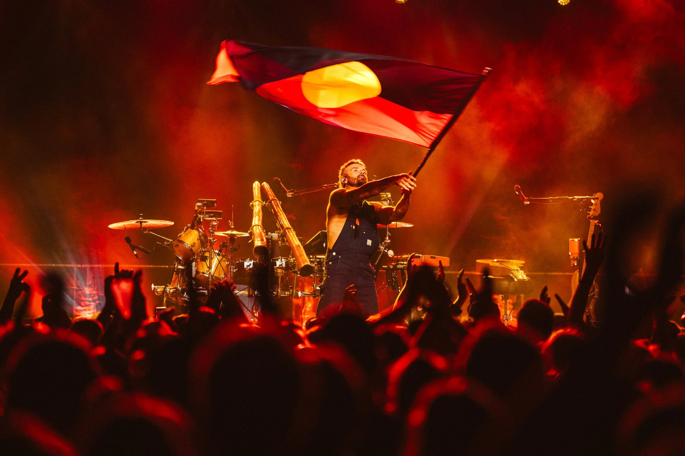 A performer on stage holding an Australian flag during a concert with a crowd in the foreground and musical instruments in the background, lit with red and orange lights.