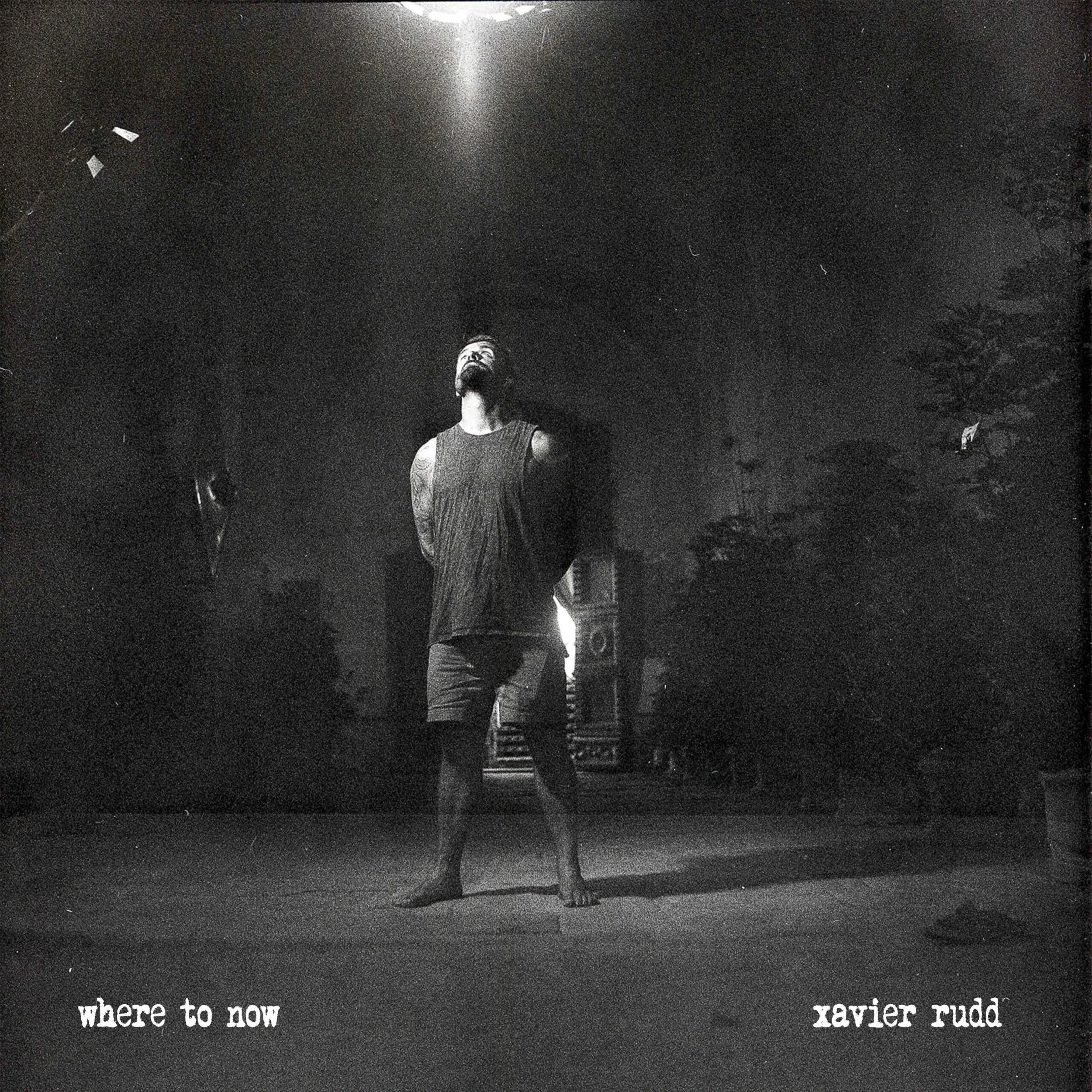 Black and white photo of a man standing on a bare stage at night, illuminated by a spotlight from above. The man is barefoot, wearing a sleeveless shirt, and facing upward with his hands behind his back. The background shows trees and a building or c