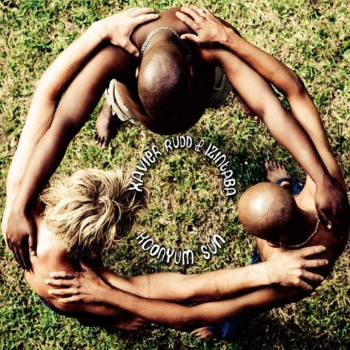Two people forming a heart shape with their bodies and arms on grass, with text reading "Xavier Rudd & Izinga Babi Sun Woonah" in the center.