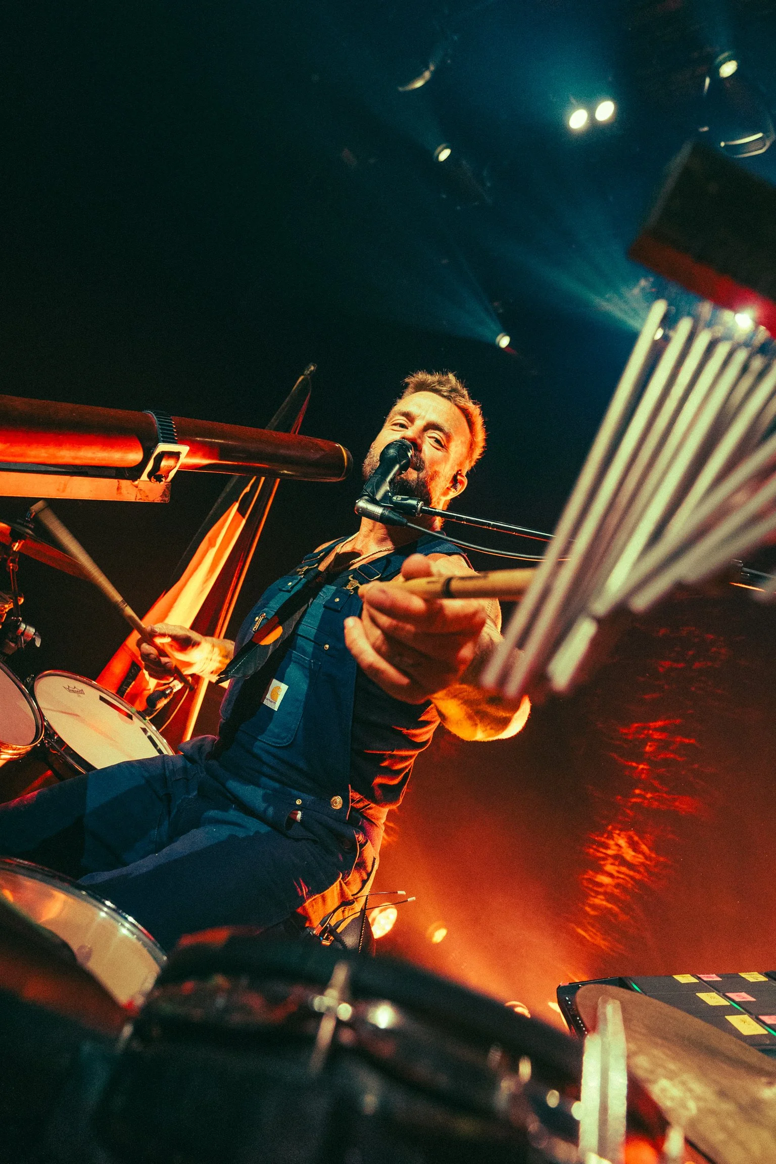 A male musician performs on stage with a drum set, microphone, and sheet music, illuminated by stage lights.