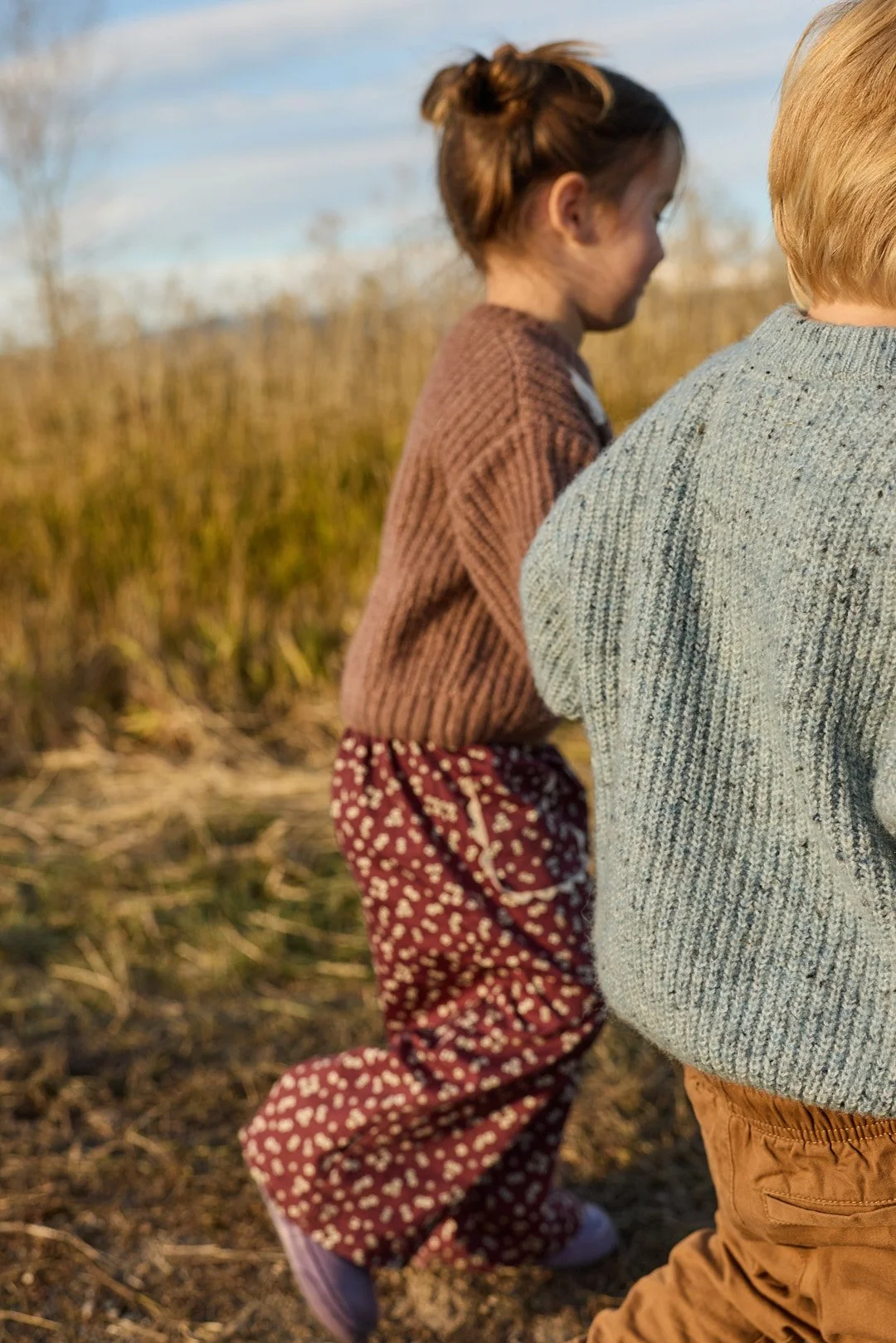 Two children outdoors, one girl in a brown sweater and patterned pants, and one boy in a gray sweater and brown pants, standing in a grassy field with trees in the background.