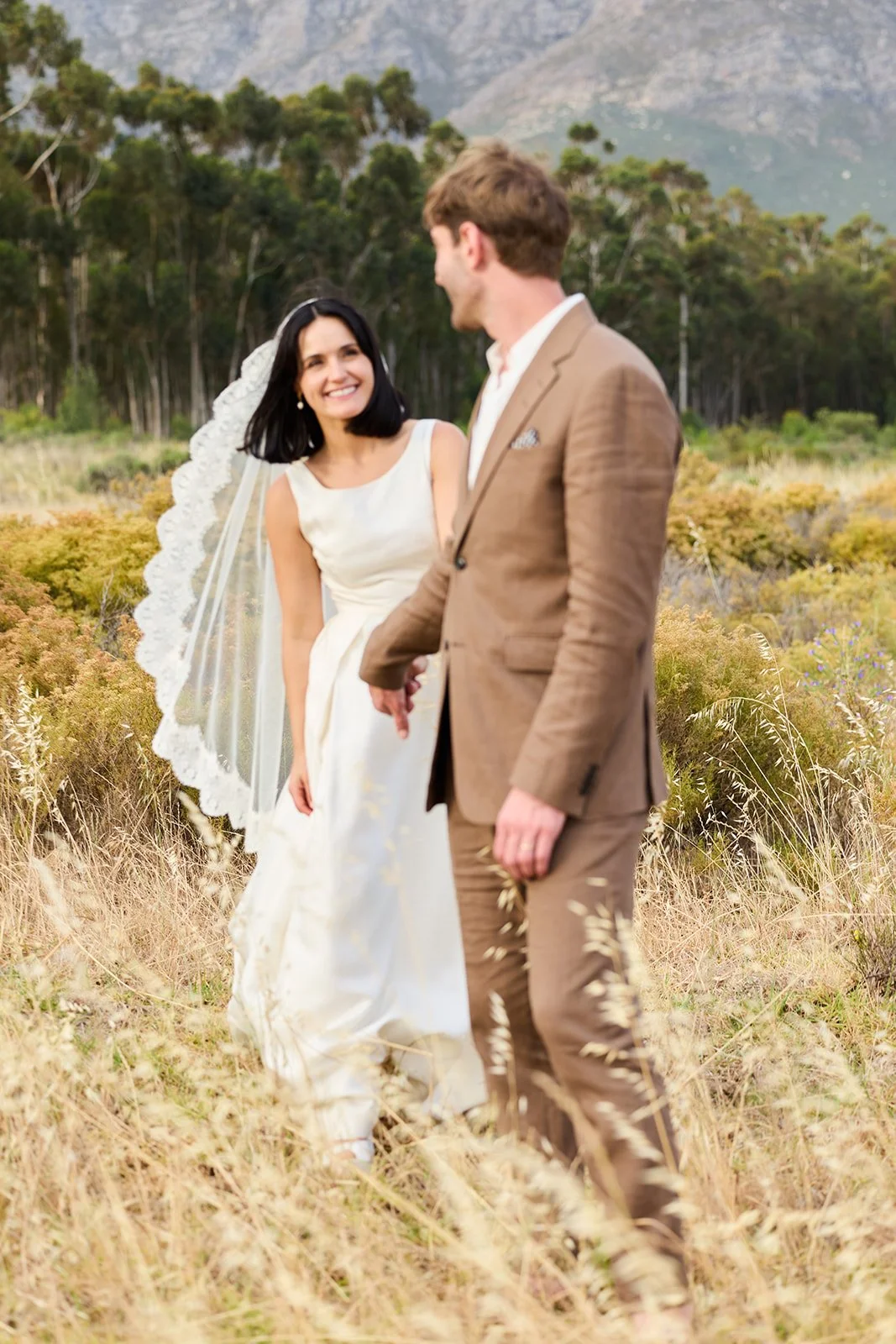 A bride and groom holding hands outdoors in a grassy field with mountains and trees in the background. The bride is smiling and wearing a white dress with a lace-edged veil, while the groom is dressed in a tan suit.