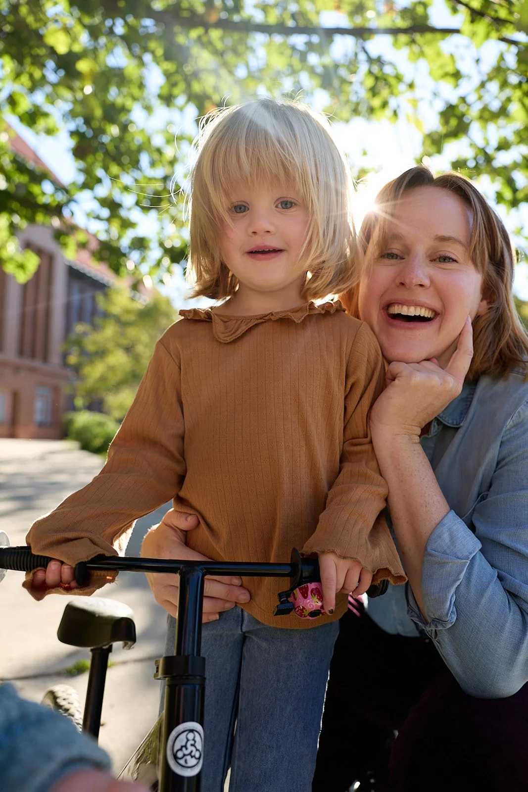 A woman and a young girl smiling outdoors on a sunny day, with the woman holding the girl and a scooter in the foreground.