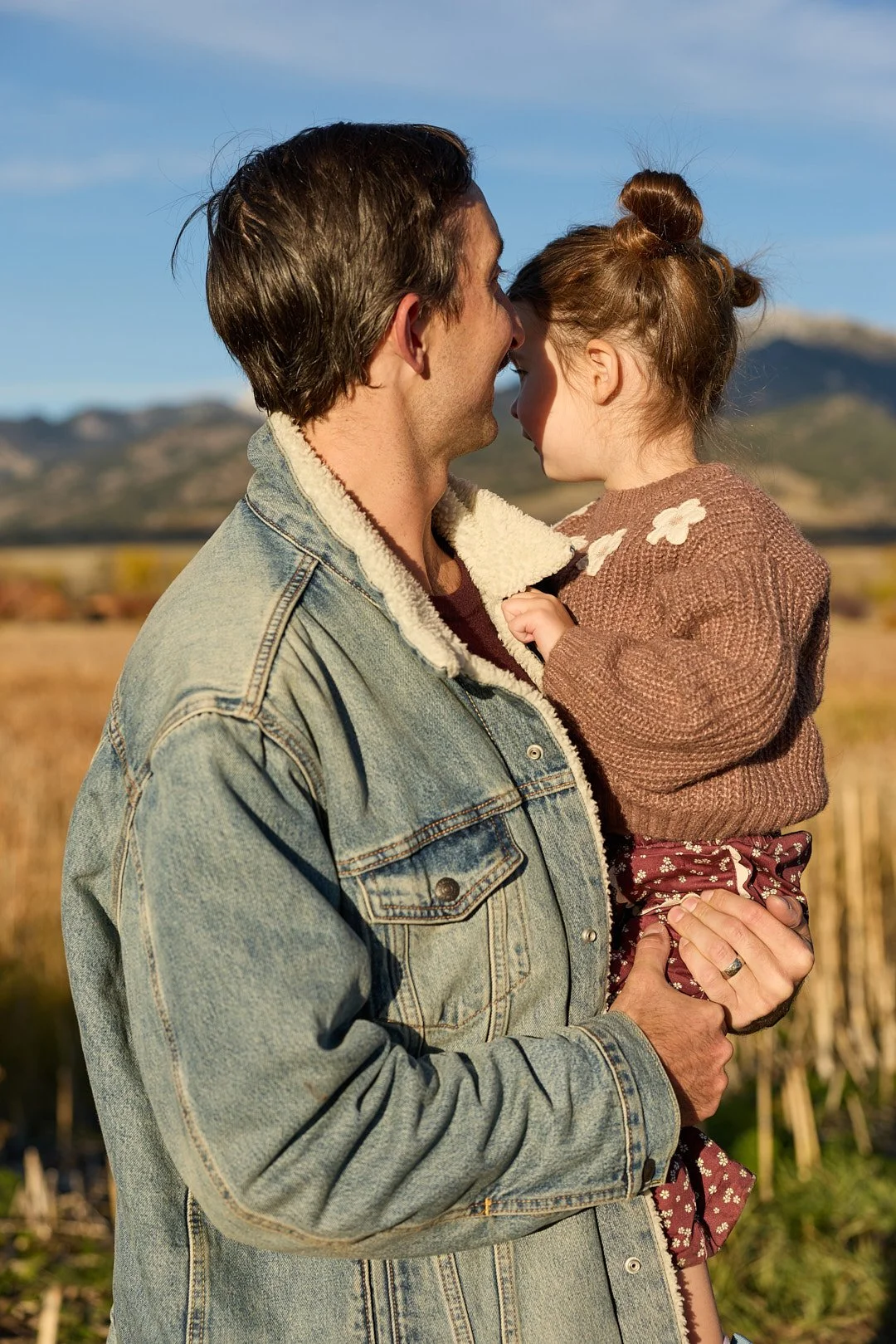 A man holding a young girl close, with their foreheads touching in an outdoor setting with mountains in the background.