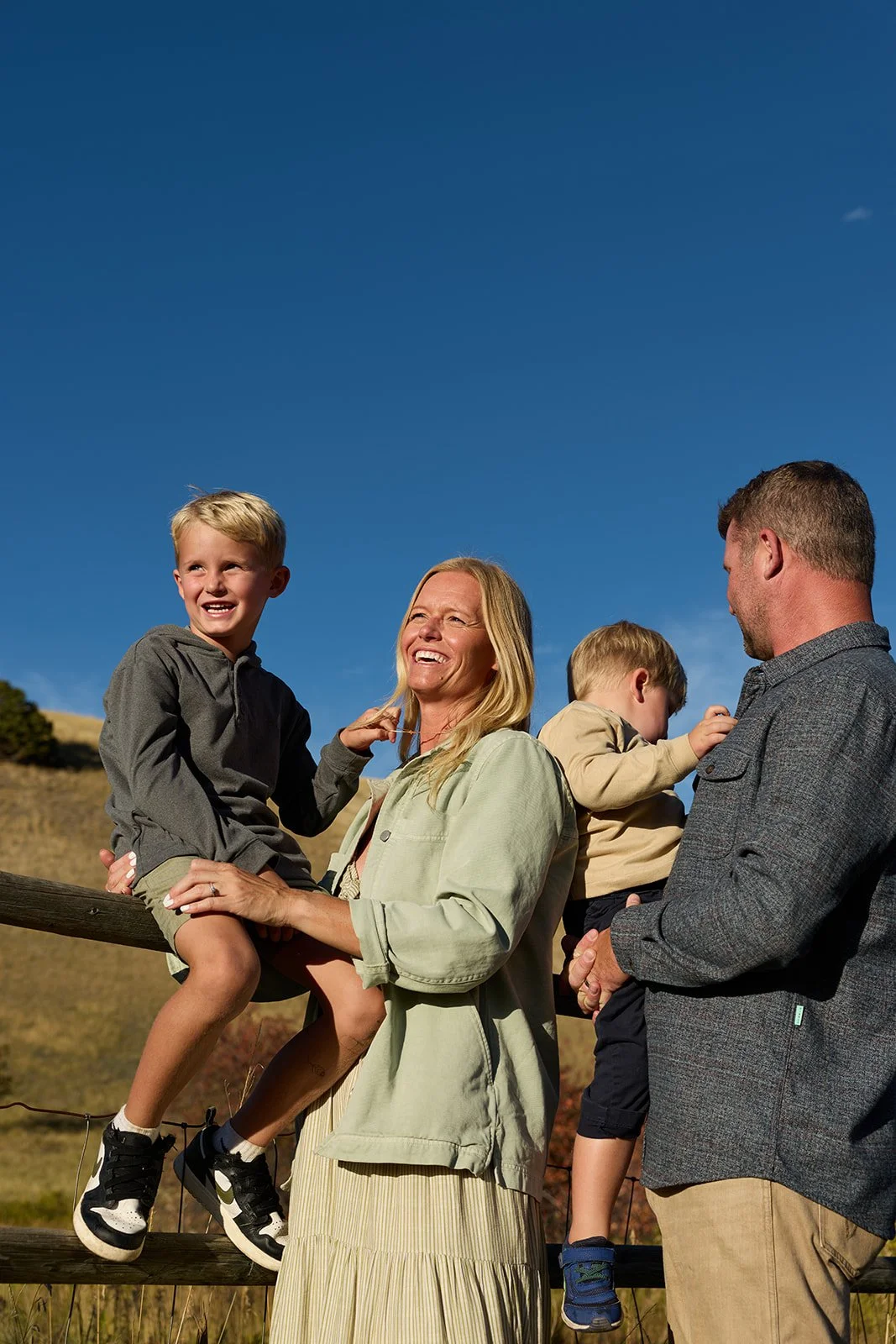 A smiling woman and two young boys outdoors on a sunny day, with a clear blue sky, standing by a wooden fence in a rural area.