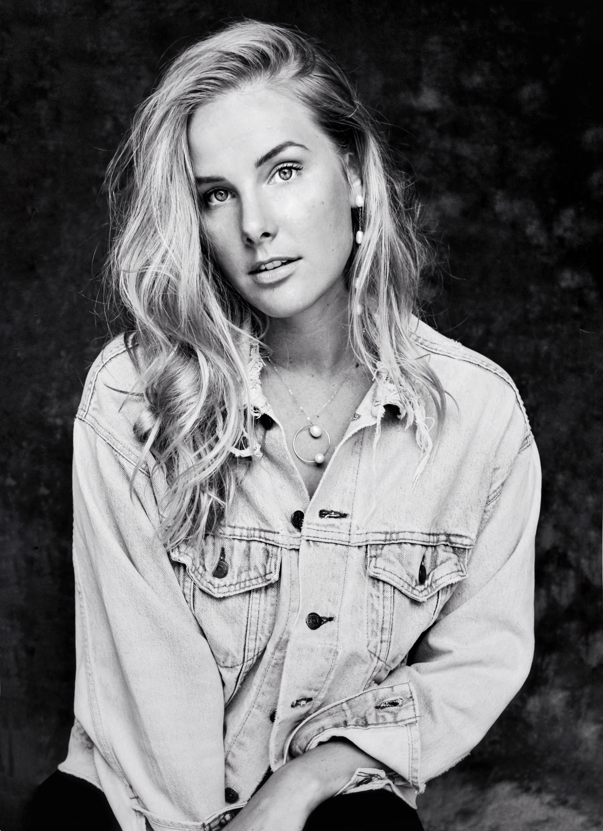Black and white portrait of a young woman with wavy hair wearing a denim jacket and layered necklaces, posing against a dark textured background.