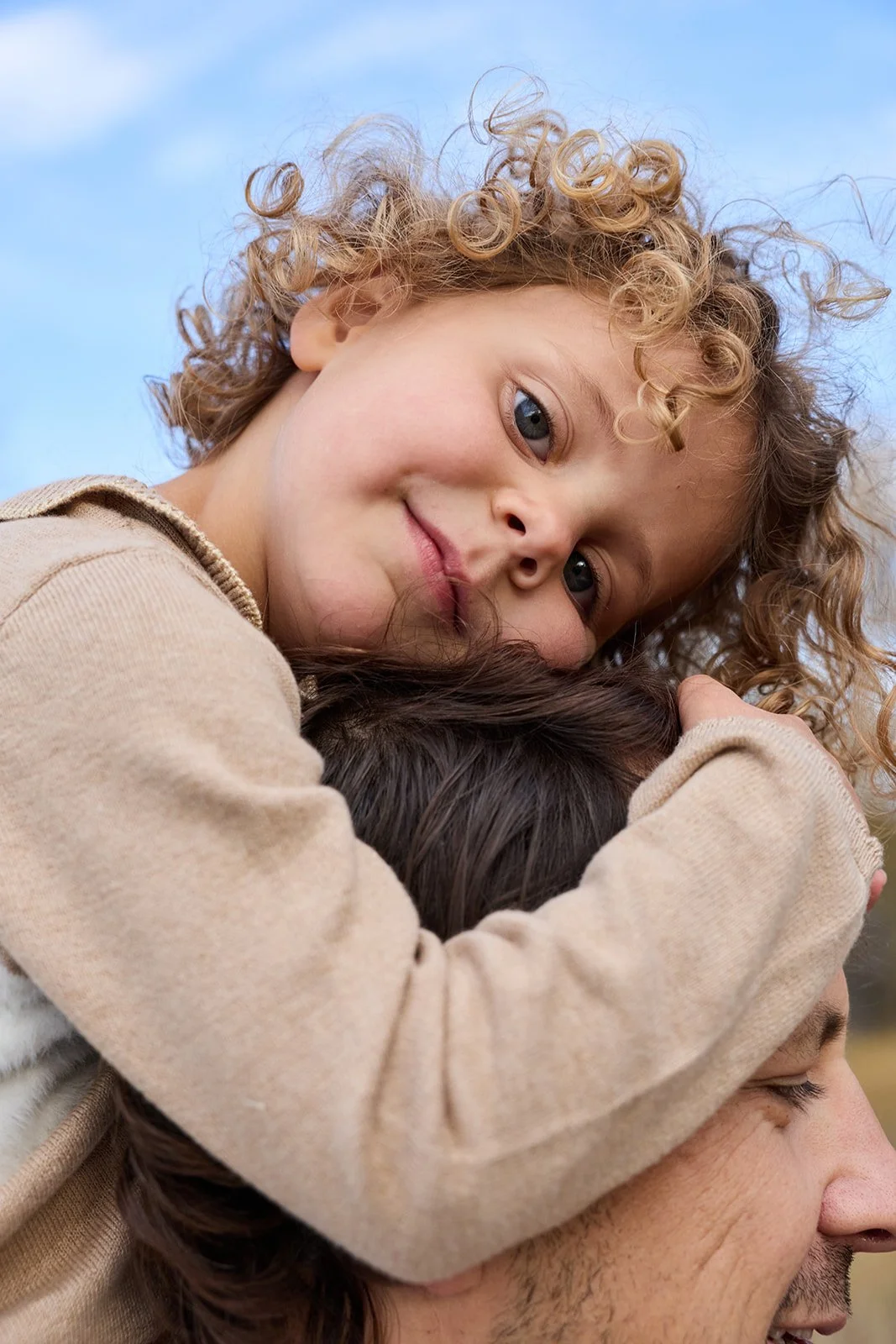 A young girl with curly blond hair hugging a man, likely her father, outdoors against a blue sky.