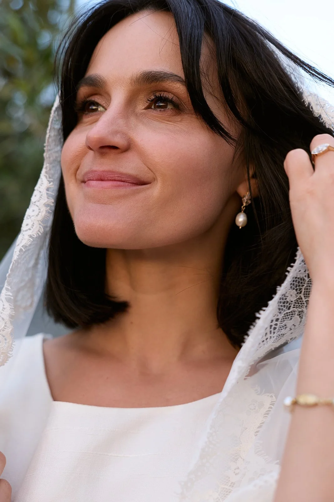 Close-up of a smiling woman with dark hair, wearing pearl earrings, a white top, and a lace veil, outdoors with blurred greenery in the background.