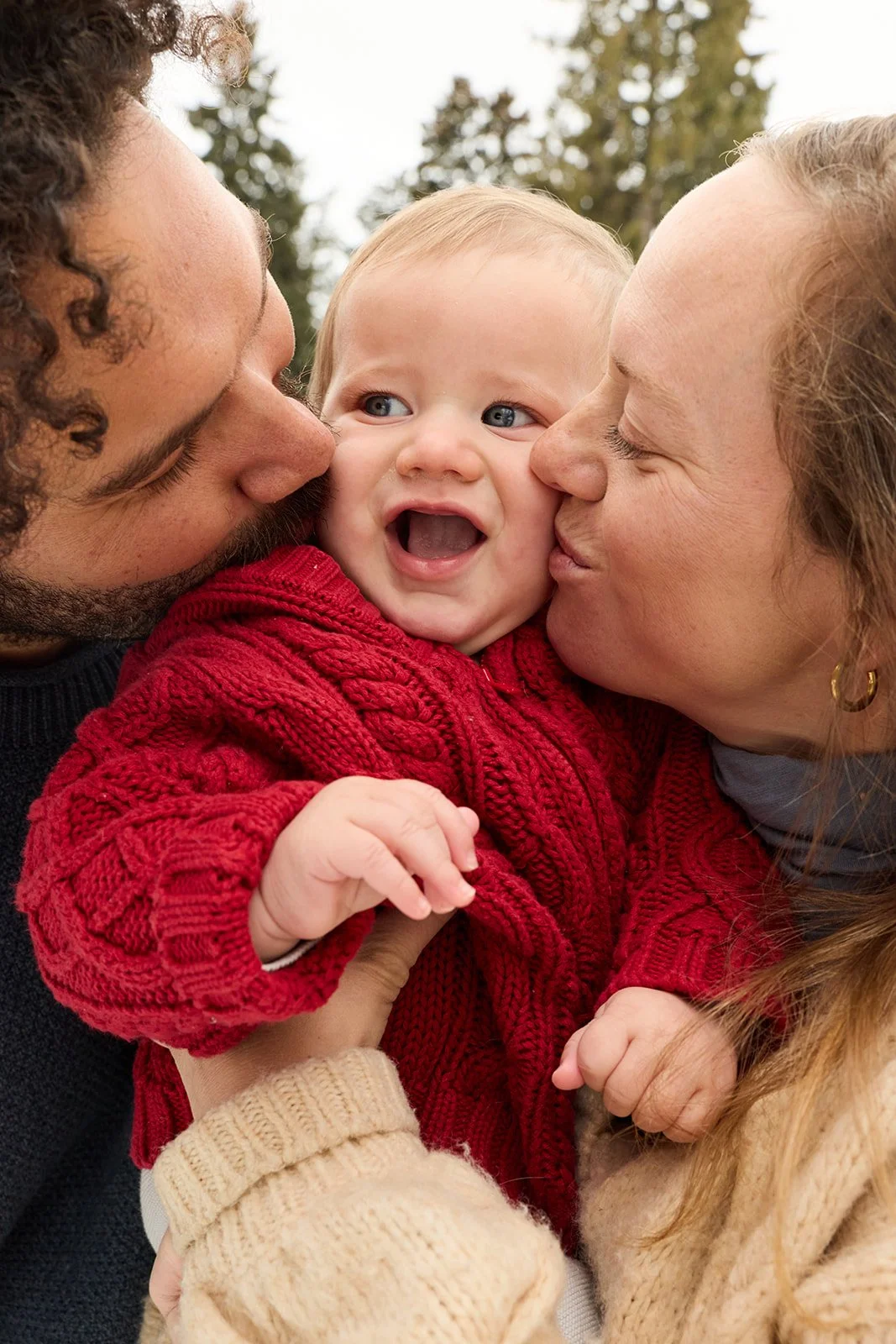 A happy baby wearing a red sweater is being kissed on each cheek by a woman and a man, all outdoors with trees in the background.