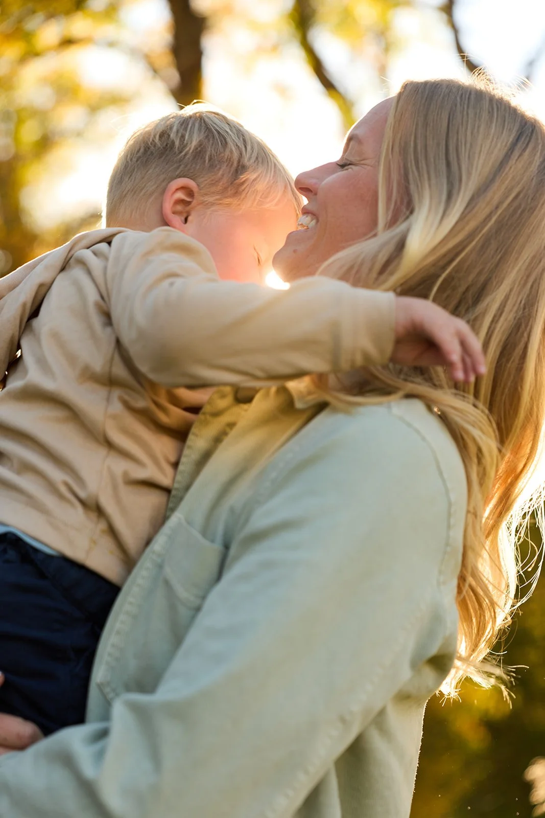 A woman holding a young child, both smiling and leaning their foreheads together outdoors during sunset with golden light, trees with autumn leaves in the background.
