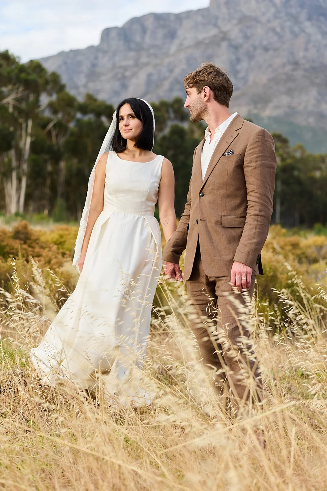 A bride and groom standing in a field of tall grass outdoors, with mountain and tree background, during their wedding.