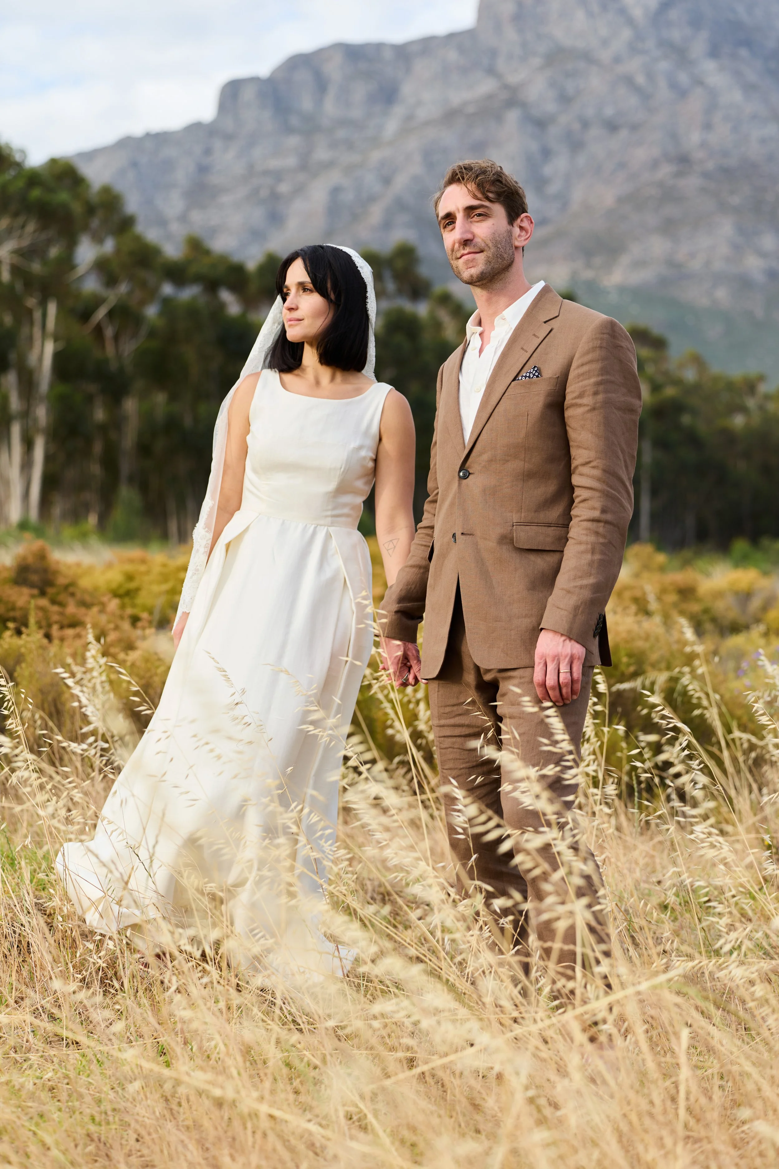 A man and woman standing in a field of tall, dry grass with mountains and trees in the background. The woman is wearing a white wedding dress with a veil, and the man is wearing a tan suit with a white shirt. They are looking off into the distance.