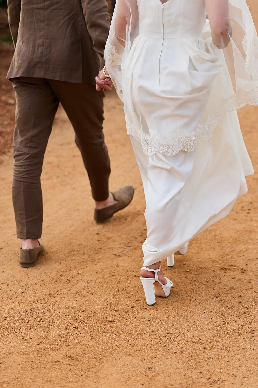 Close-up of bride and groom walking on dirt path, holding hands, bride in white wedding dress with lace, wearing white high heel shoes, groom in brown suit trousers and brown shoes.