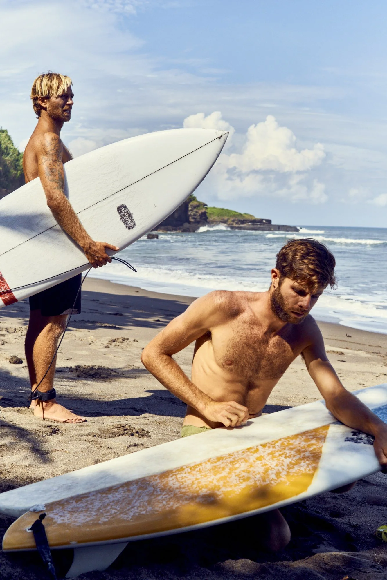 Two men on a beach preparing surfboards, with one holding a surfboard and the other kneeling on the sand, with ocean waves and cloudy sky in the background.