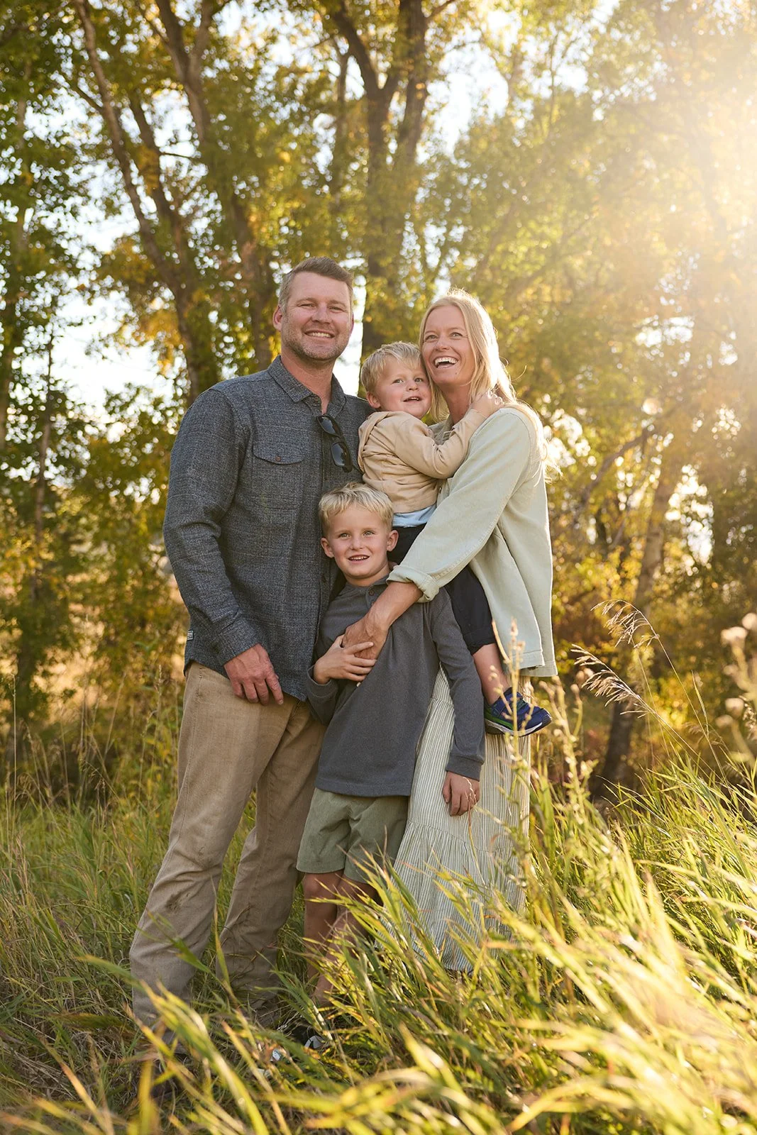 Family of four outdoors in a grassy area, sun shining through trees in the background, smiling and embracing each other.