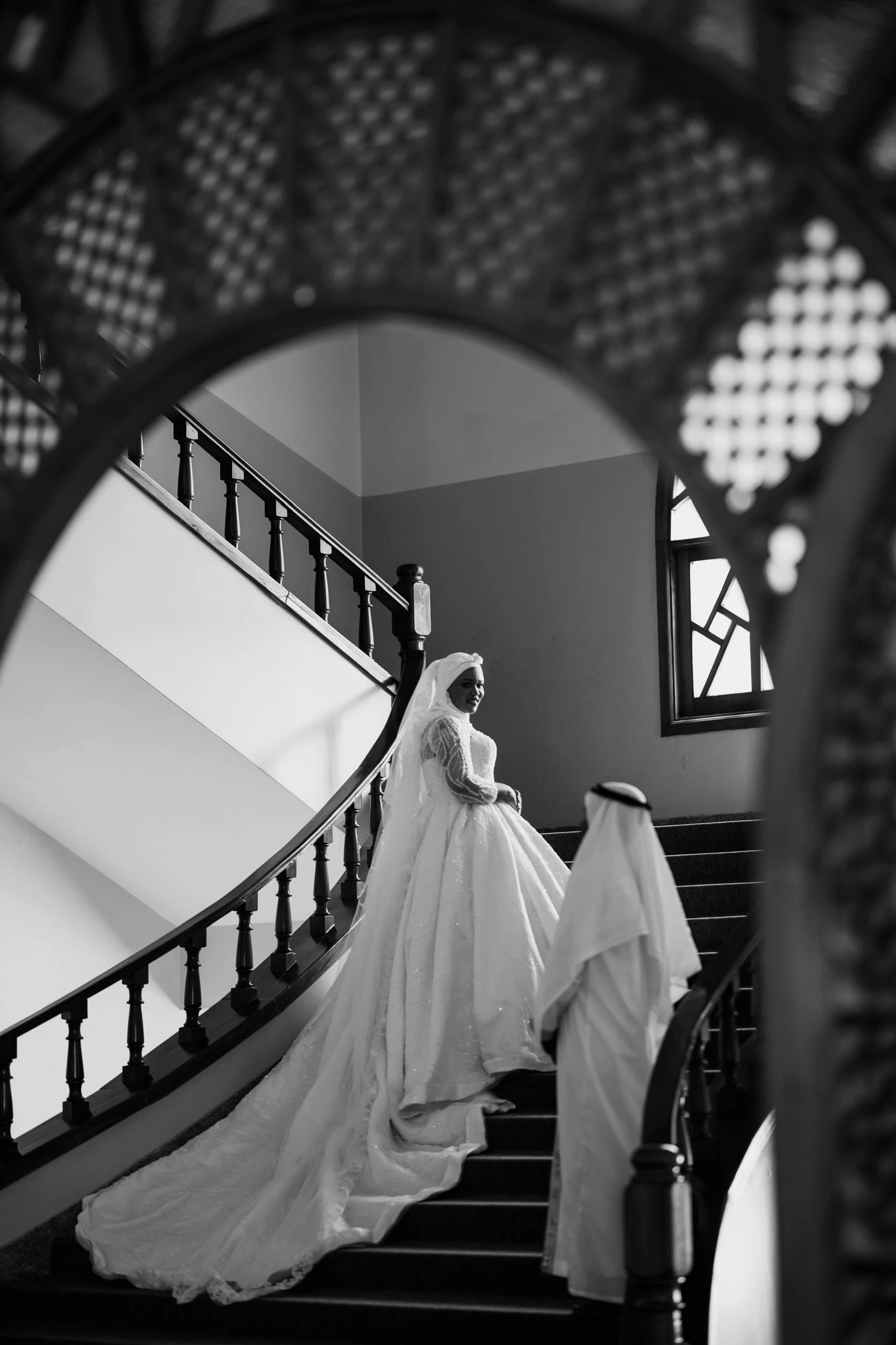 A bride in a wedding dress standing on a curved staircase, seen through an intricate decorative frame with a window in the background.