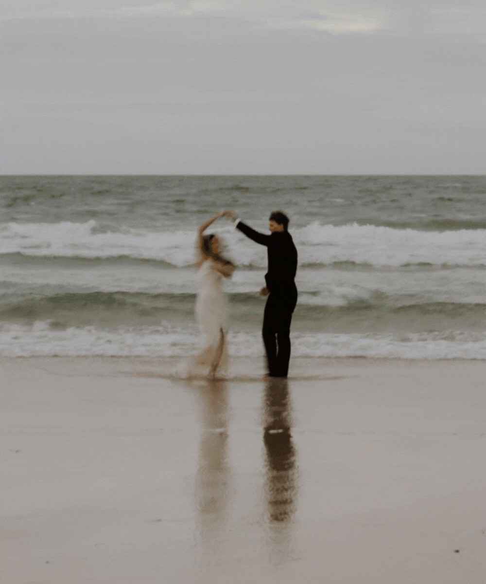 A couple dancing on the beach near the ocean, with waves in the background.