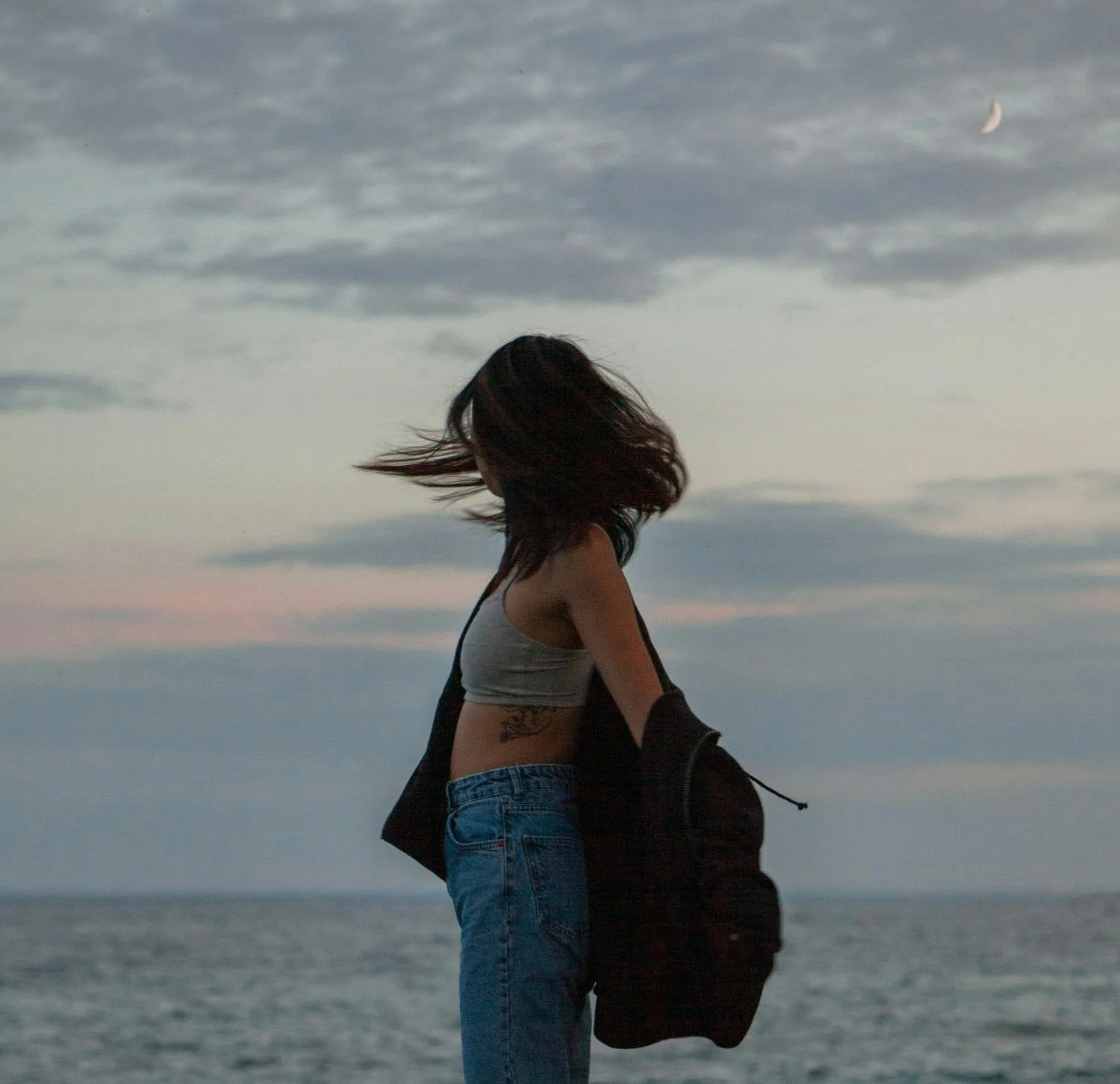 A woman standing outdoors by the water during dusk, with her hair blowing in the wind, wearing a crop top, jeans, and a jacket draped over her arm, under a cloudy sky with a crescent moon.