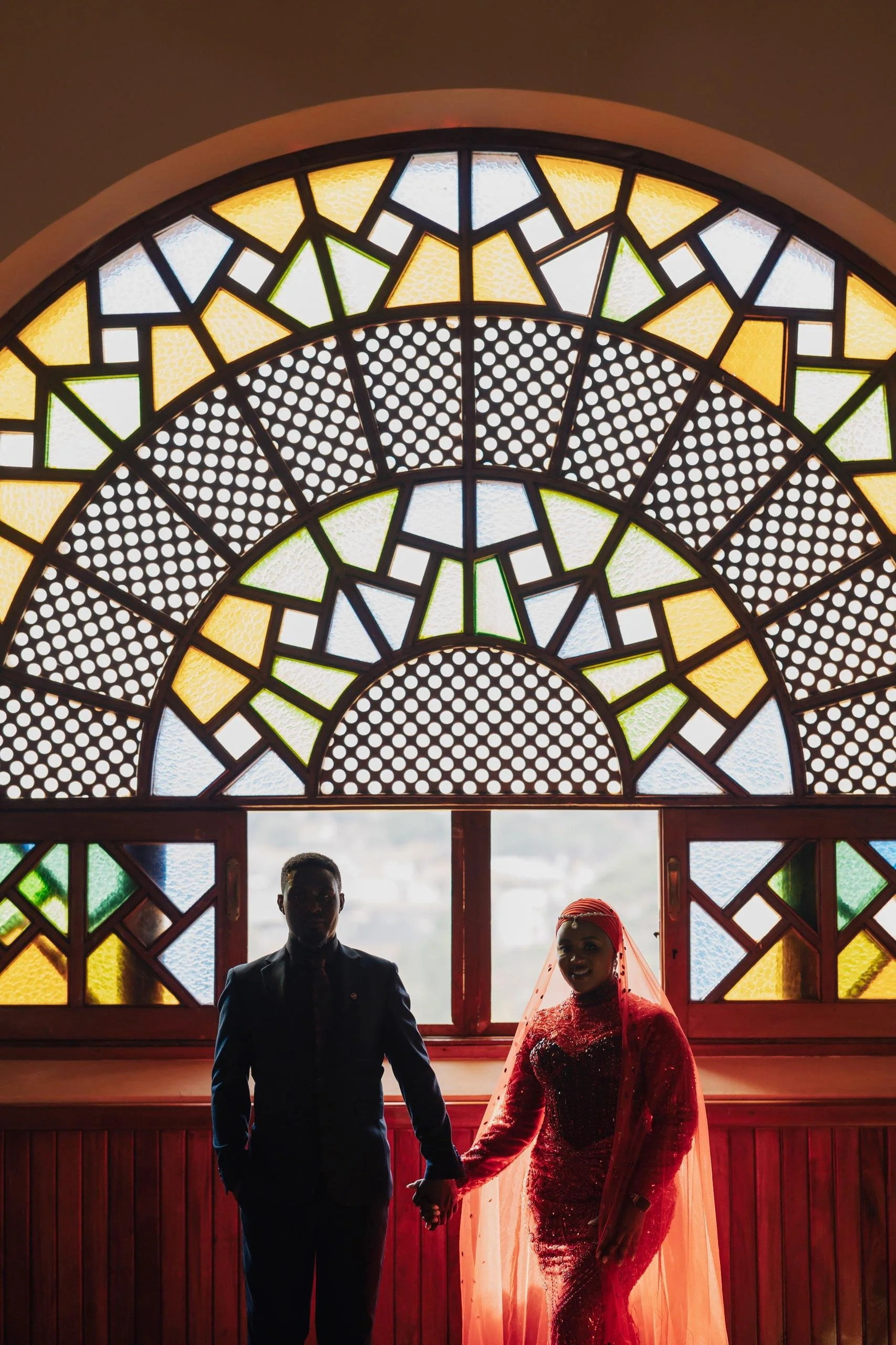 A couple in formal attire holding hands, standing in front of a large stained glass window with geometric patterns and yellow, green, blue, and white colors.