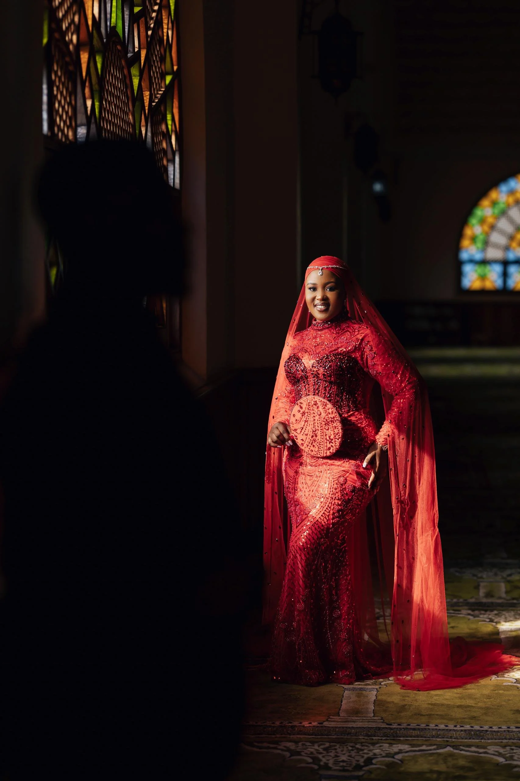 A woman in a red, intricately designed dress with a matching veil stands inside a church, illuminated by natural light from stained glass windows.