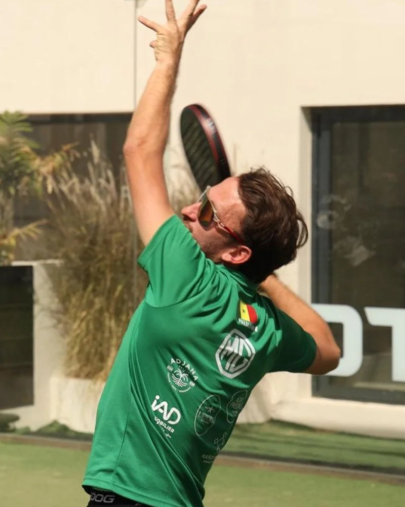 Un homme joue au tennis, portant une chemise verte, des lunettes de soleil et un bandeau, en train de faire un mouvement de service.