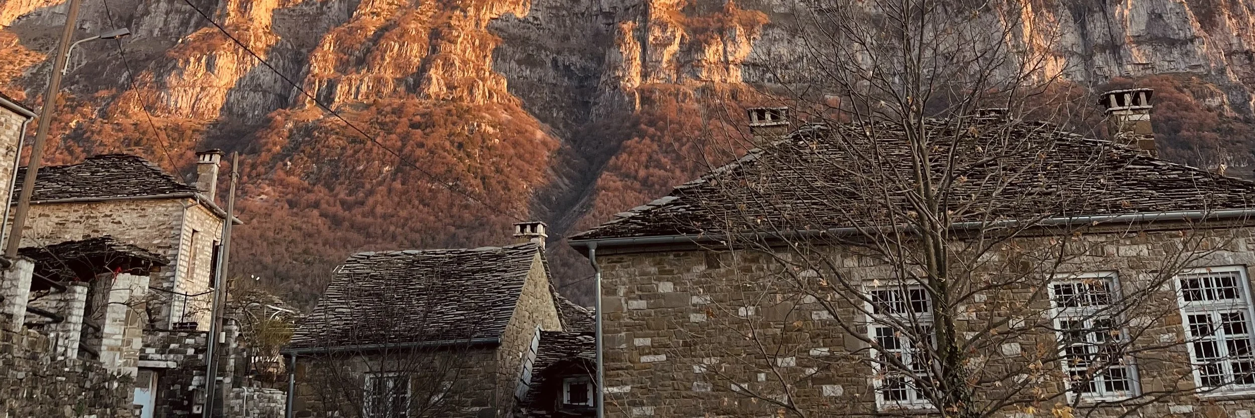 Stone houses with slate roofs in a mountain village, with leafless trees and a backdrop of rugged, sunlit mountain peaks.
