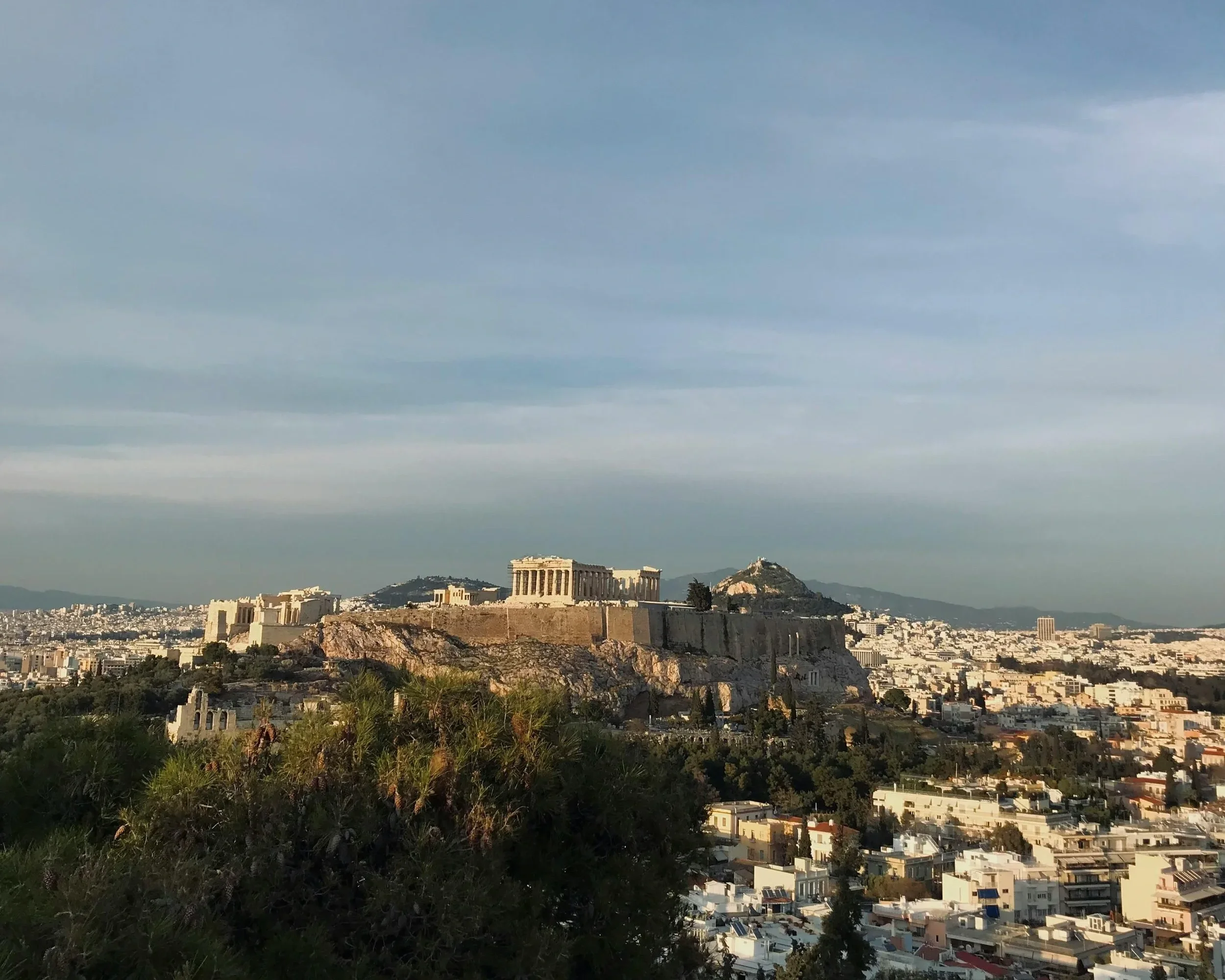 View of the ancient Acropolis in Athens, Greece, with the Parthenon temple on a hill, under a partly cloudy sky at sunset, surrounded by city buildings and lush greenery.