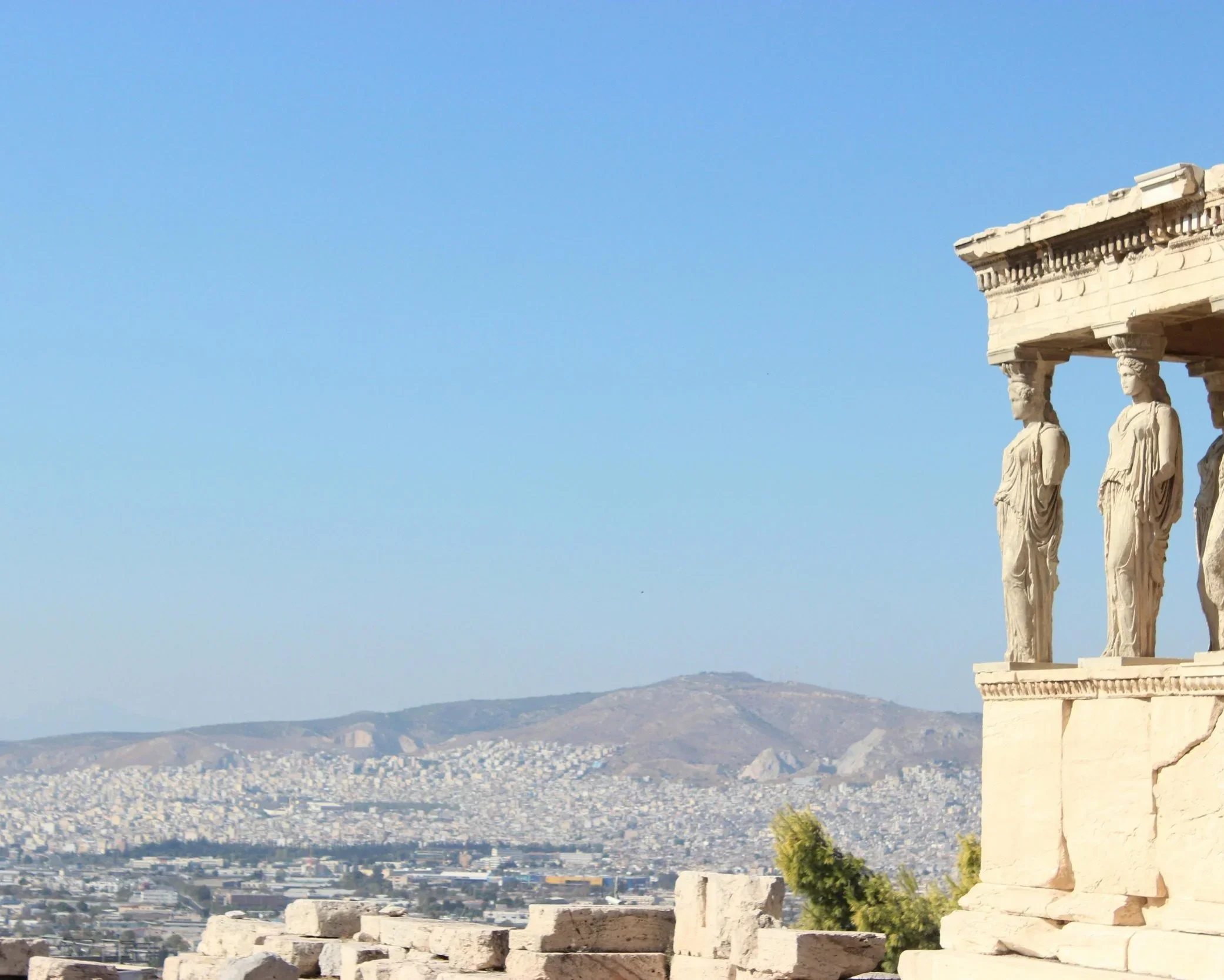 Ancient Greek temple ruins with columns and sculptures on a hill overlooking a city, with hills and mountains in the background.
